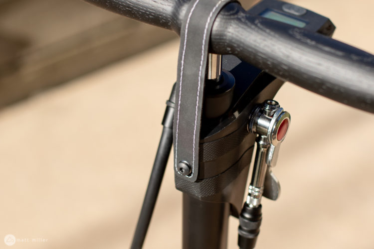 Close-up view of a bicycle handlebars and stem, featuring a digital display and a valve for air adjustment or inflation. The handlebars exhibit a textured black finish, while the stem is designed with a sleek, modern look. Sunlight highlights the details of the materials and stitching.