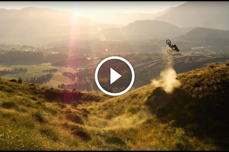A mountain biker performing a backflip over a grassy hill against a scenic landscape, with rolling hills and a sunset illuminating the background. Dust is kicked up from the ground as the biker showcases an impressive aerial trick.