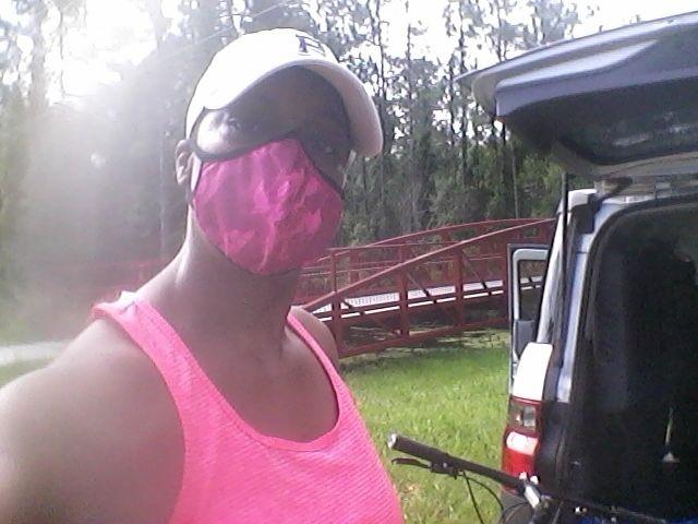 A person wearing a bright pink tank top and a pink mask stands outdoors, near a vehicle with its trunk open. In the background, a red pedestrian bridge and greenery are visible, indicating a natural setting. The individual is also wearing a white cap. Hidden Falls Regional Park mountain bike trail.