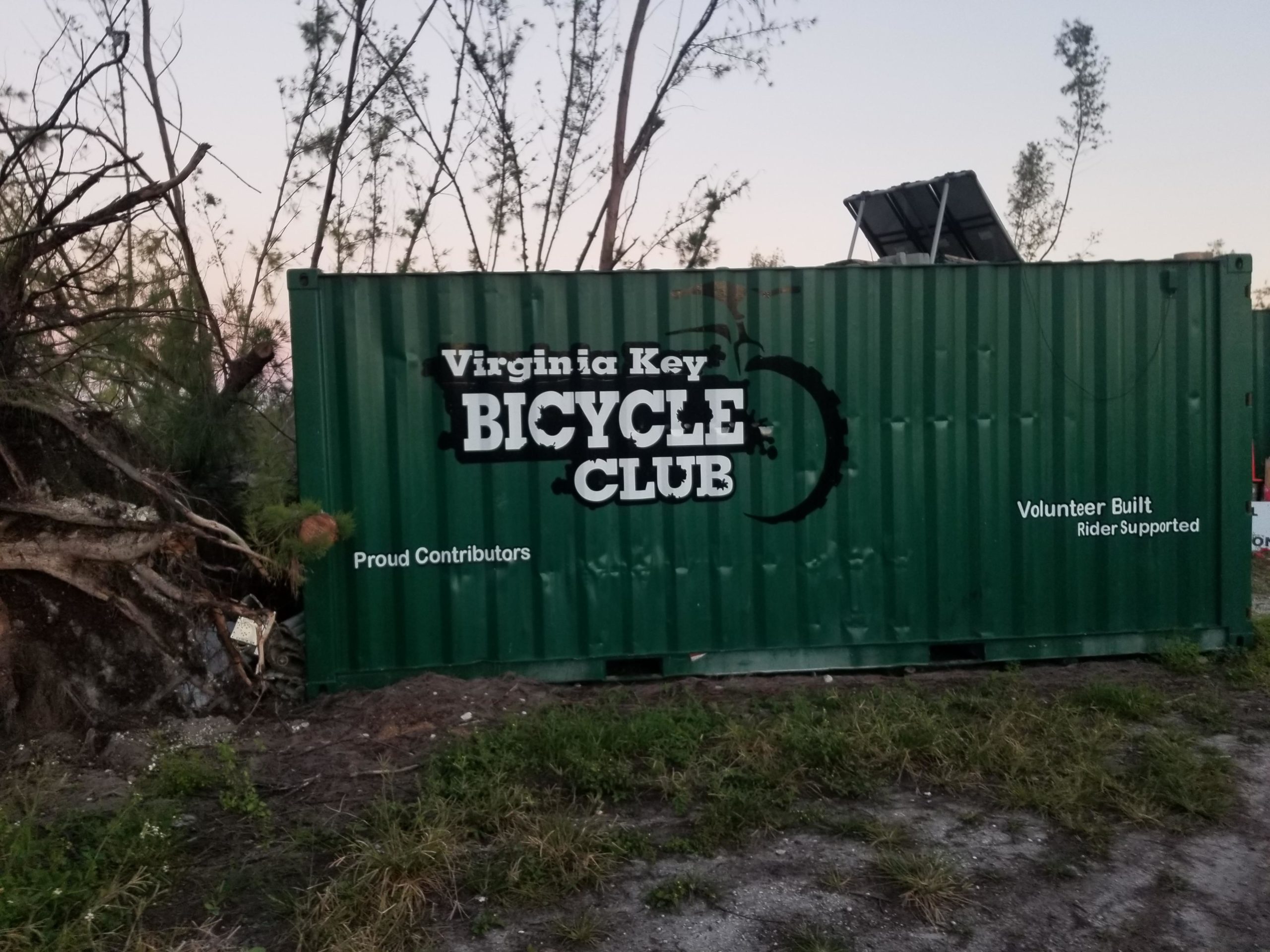 A green shipping container with the text "Virginia Key BICYCLE CLUB" prominently displayed. Additional text includes "Proud Contributors" and "Volunteer Built, Rider Supported." The background features trees and vegetation. Virginia Key North Point mountain bike trail.