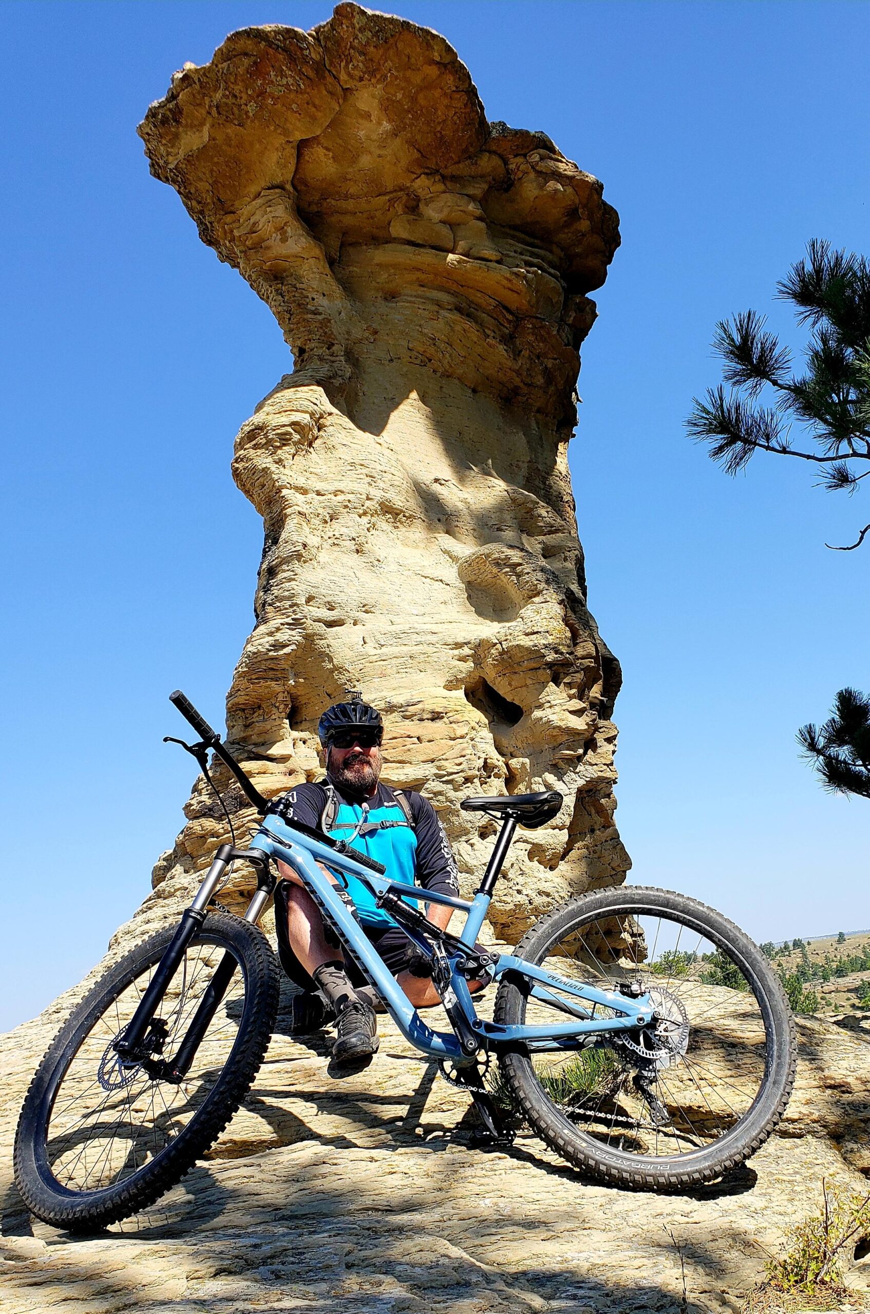 Specialized Stumpjumper: A mountain biker sitting on a rock formation, with a blue bike resting beside him. The scene features a clear blue sky with no clouds, showcasing a beautiful outdoor landscape. The rock formation is tall and unique in shape, while pine trees are visible in the background. The cyclist is wearing a helmet and a blue and black cycling jersey, smiling at the camera.