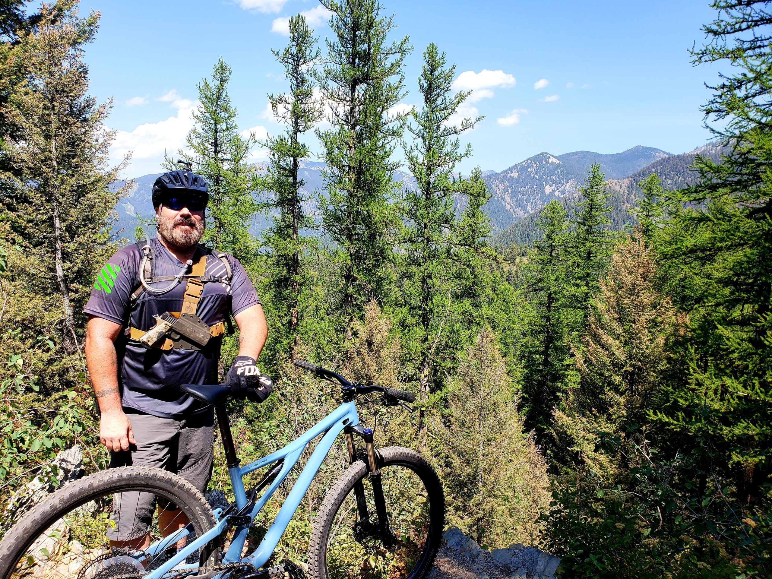 A mountain biker standing next to a blue mountain bike on a forested trail, surrounded by tall green trees and mountains in the background under a clear blue sky. The biker is wearing a helmet, sunglasses, and a padded shirt, with a tool belt around their waist. Mt Helena Ridge mountain bike trail.