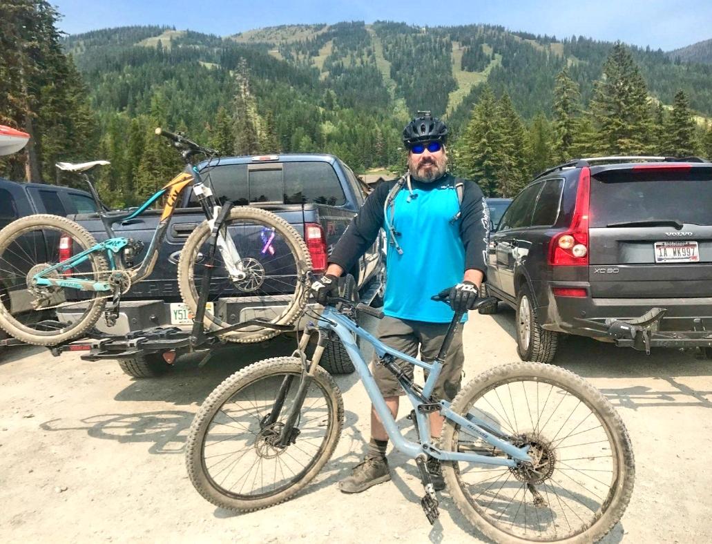 A man wearing a blue shirt and black sunglasses stands next to a parked vehicle, holding a mountain bike. A second bike is secured on a rack behind him. The background features green hills and trees, indicating a mountainous outdoor setting, with a clear blue sky overhead. Big Sky Resort mountain bike trail.