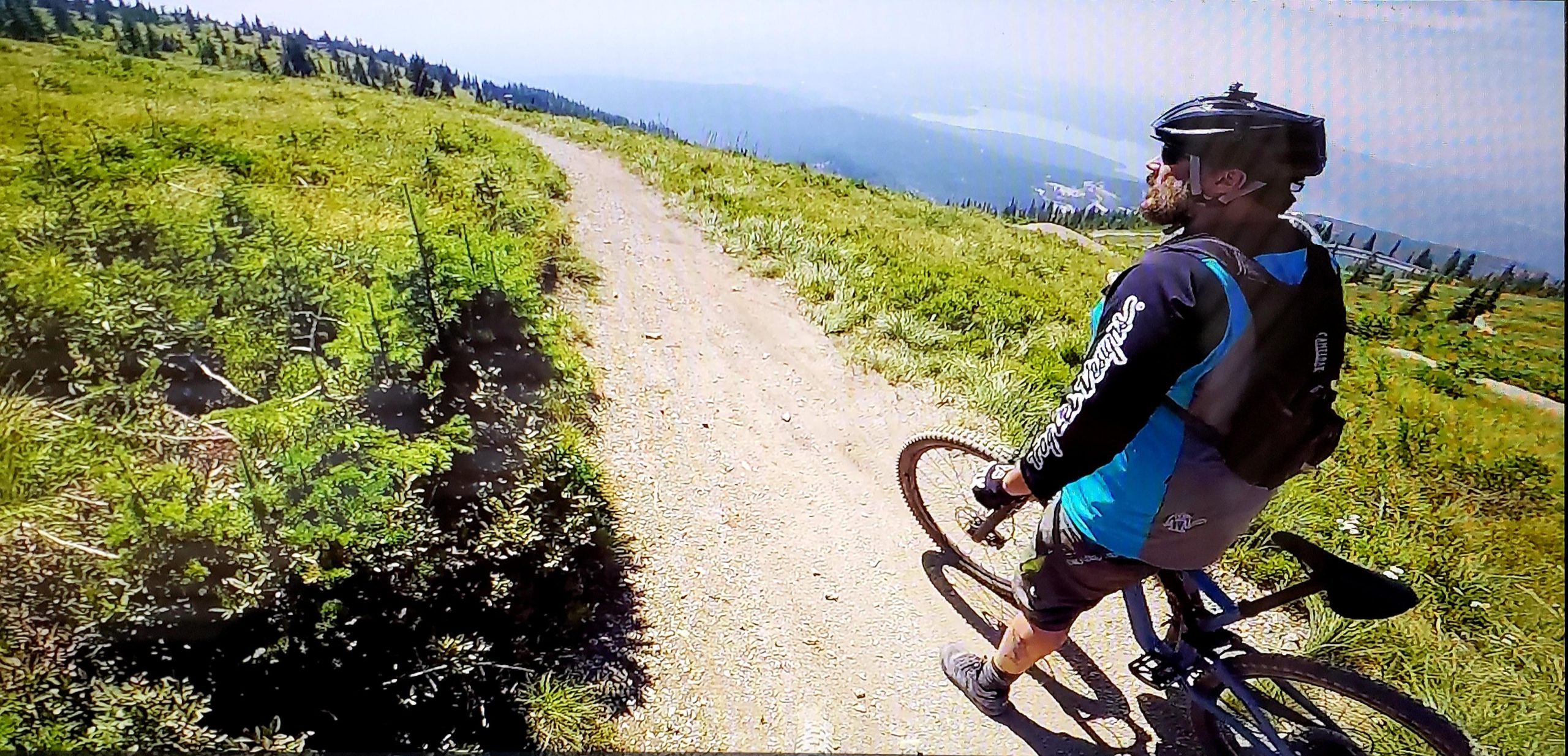 A cyclist stands beside a mountain bike on a dirt path surrounded by green vegetation and trees, with a panoramic view of hills and a distant landscape in the background. The cyclist, wearing a helmet and a blue and black cycling outfit, is looking towards the scenery. Whitefish Mountain Resort mountain bike trail.