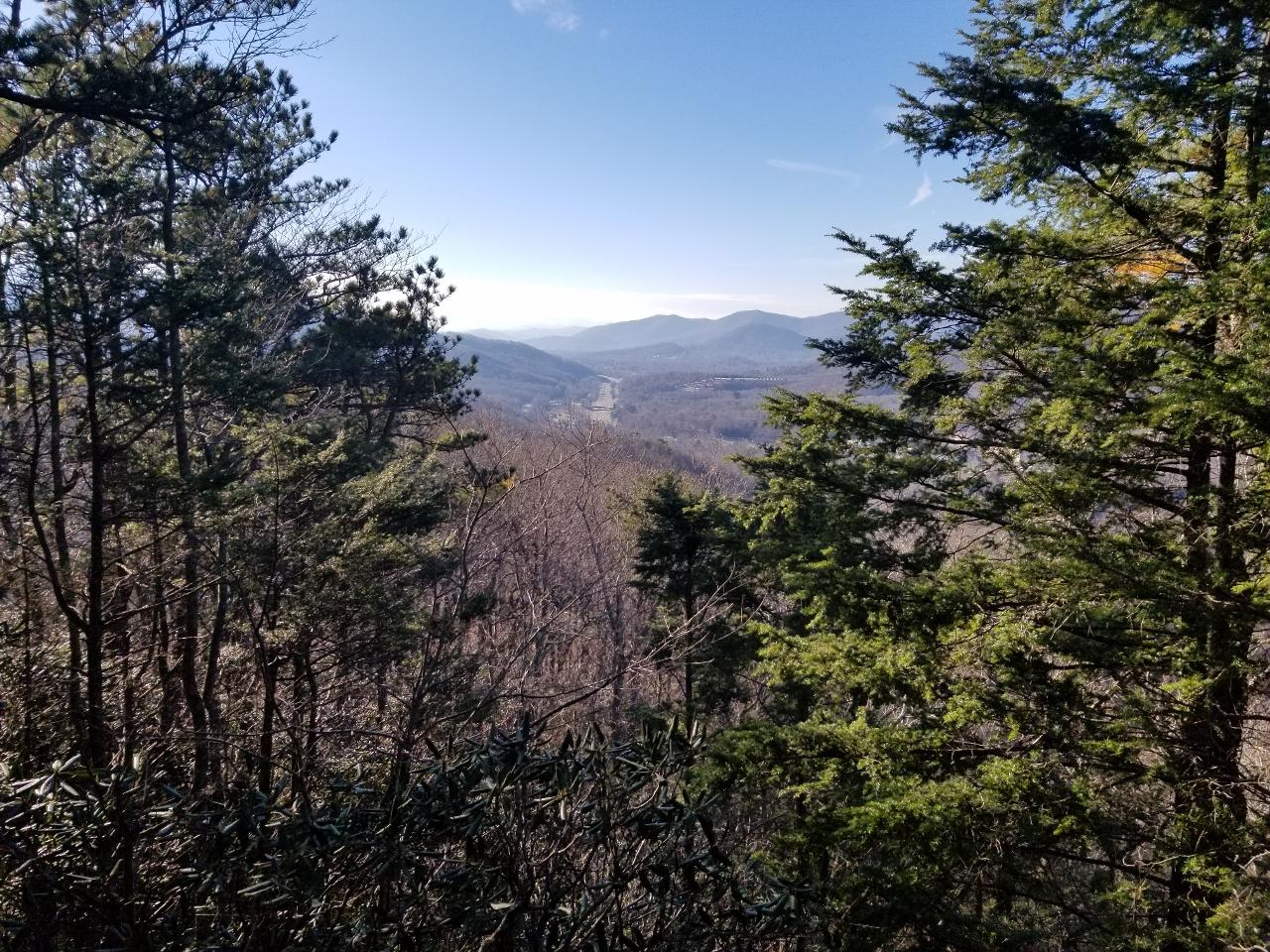 A scenic view of rolling mountains in the distance, framed by evergreen trees in the foreground, under a clear blue sky. The landscape features a winding road and bare trees, indicative of late autumn or winter. Kitsuma mountain bike trail.
