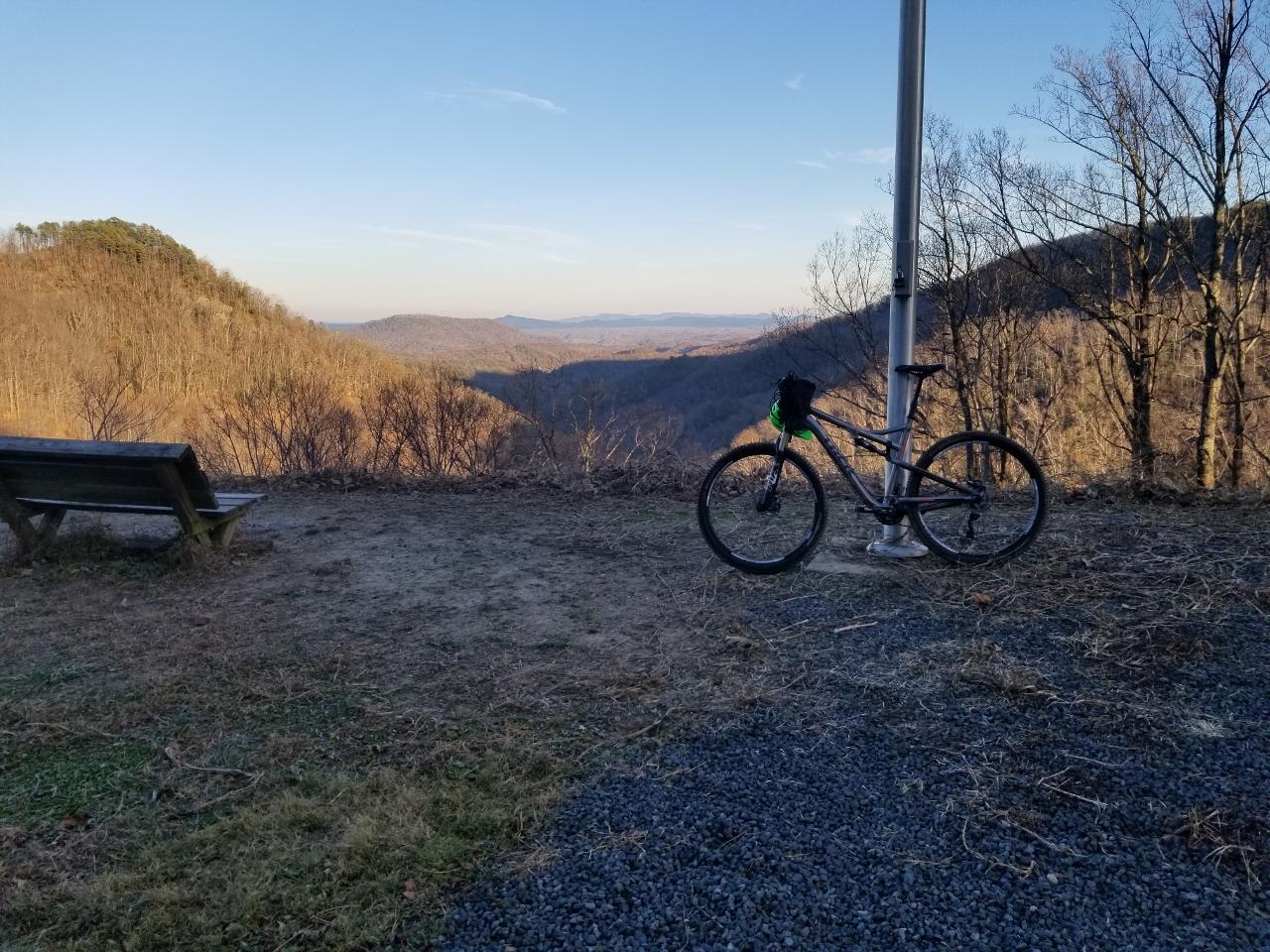 A mountain bike parked next to a flagpole, with a scenic view of rolling hills and valleys in the background. A wooden bench is visible nearby, and the landscape shows a mix of bare trees and distant mountains under a clear sky. Kitsuma mountain bike trail.