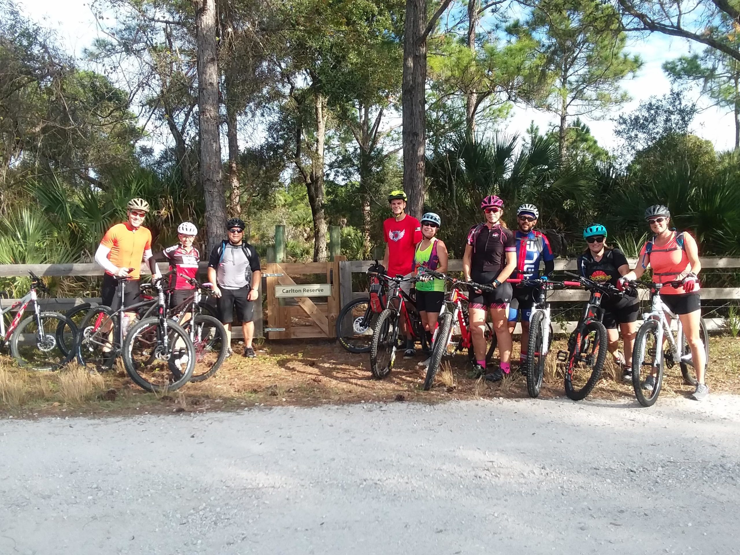 A group of eight cyclists, wearing colorful biking gear and helmets, pose for a photo at the entrance of Carlton Reserve. They are standing beside their mountain bikes on a dirt path surrounded by trees and greenery. The background features a wooden fence and a sign indicating the entrance to Carlton Reserve. Carlton Preserve mountain bike trail.