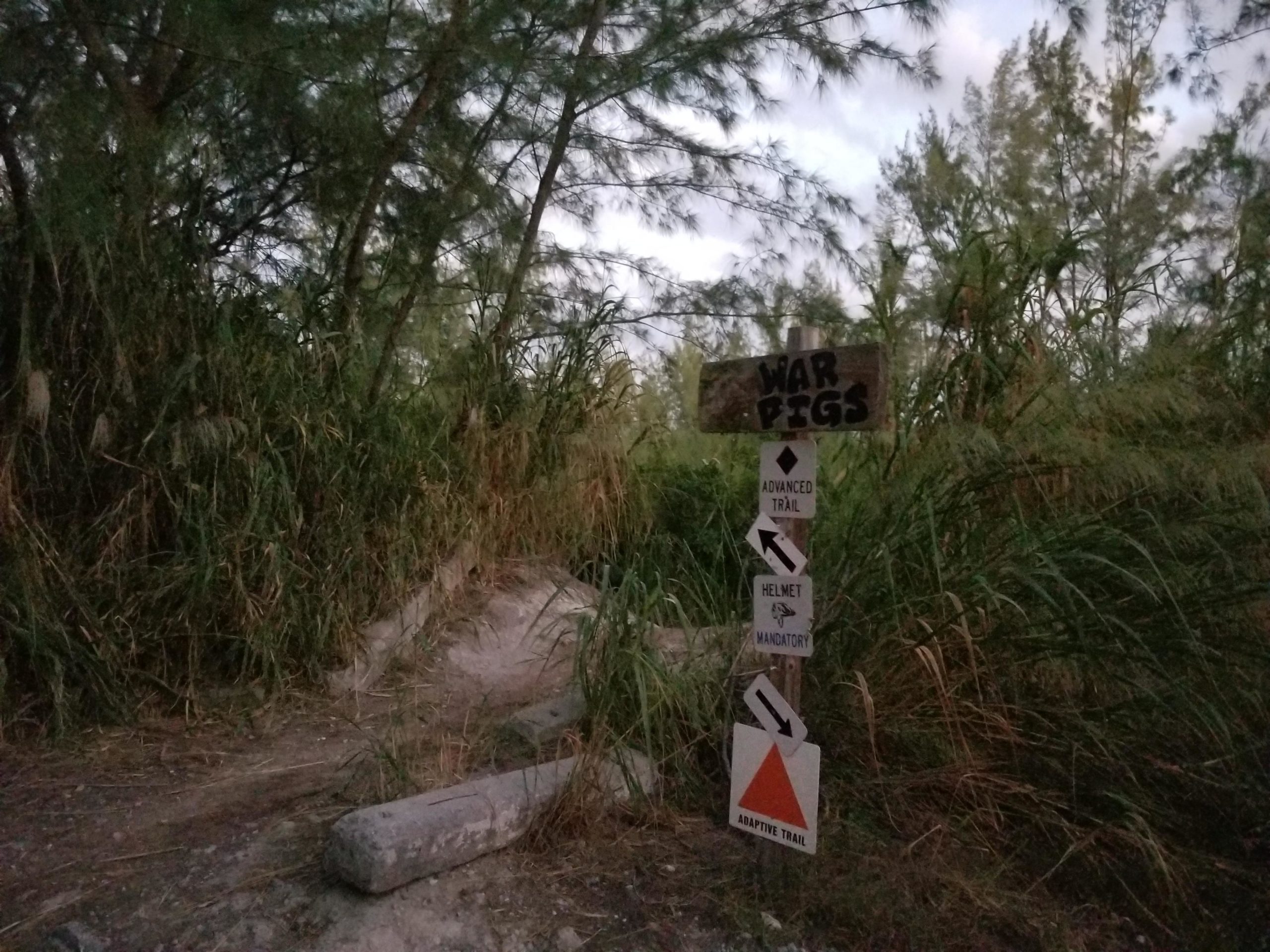 A weathered wooden sign labeled "WAR PIGS" stands amidst tall grass and trees at the entrance to a dirt path, with additional signs indicating trail difficulty and safety requirements, including "ADVANCED TRAIL," "HELMET MANDATORY," and "ADAPTIVE TRAIL." The scene is dimly lit, suggesting twilight. Virginia Key North Point mountain bike trail.