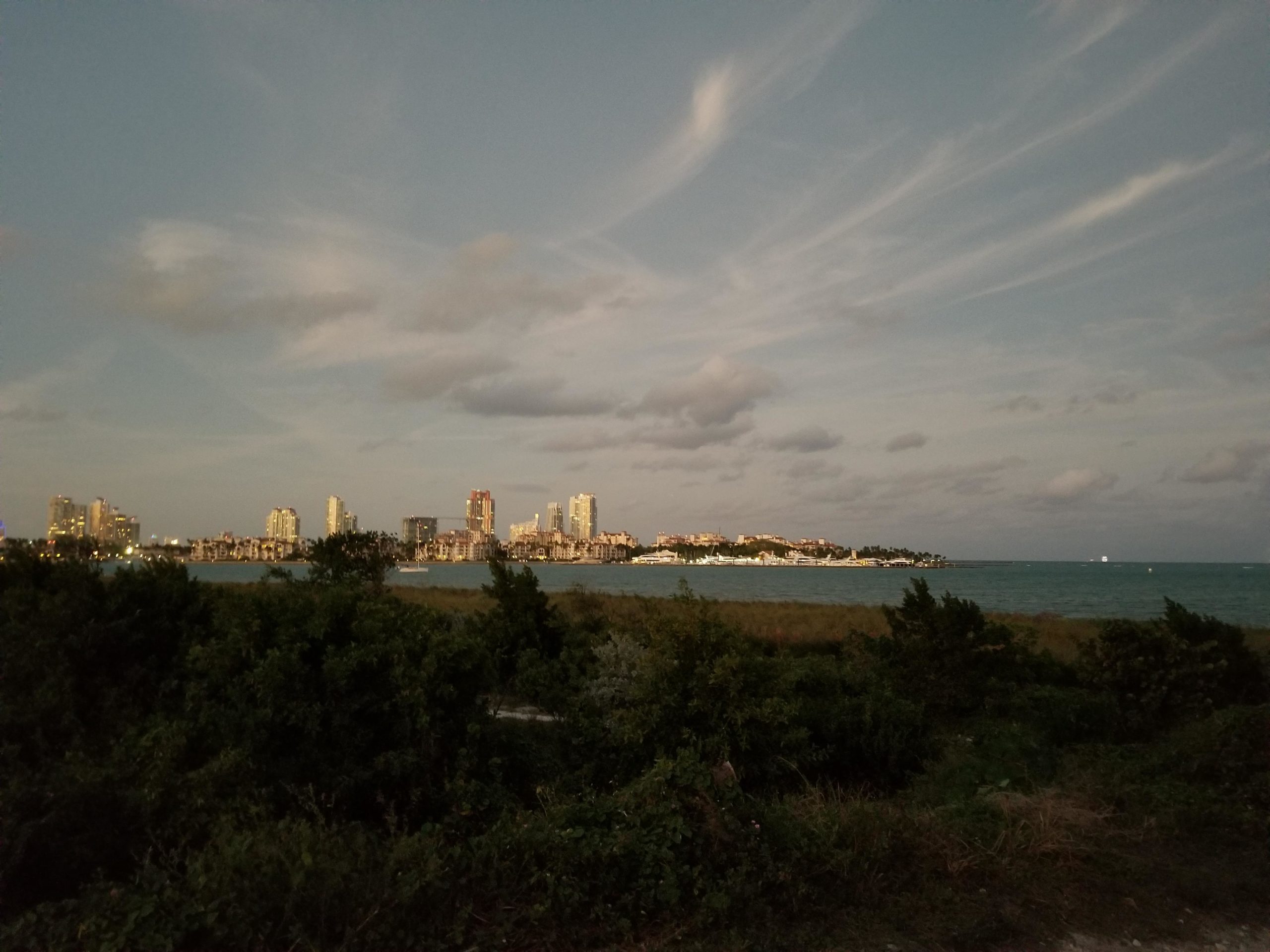 City skyline at dusk viewed from a coastal area, featuring illuminated buildings and a calm ocean. The sky is filled with clouds, and a small boat is visible on the water, surrounded by lush greenery in the foreground. Virginia Key North Point mountain bike trail.