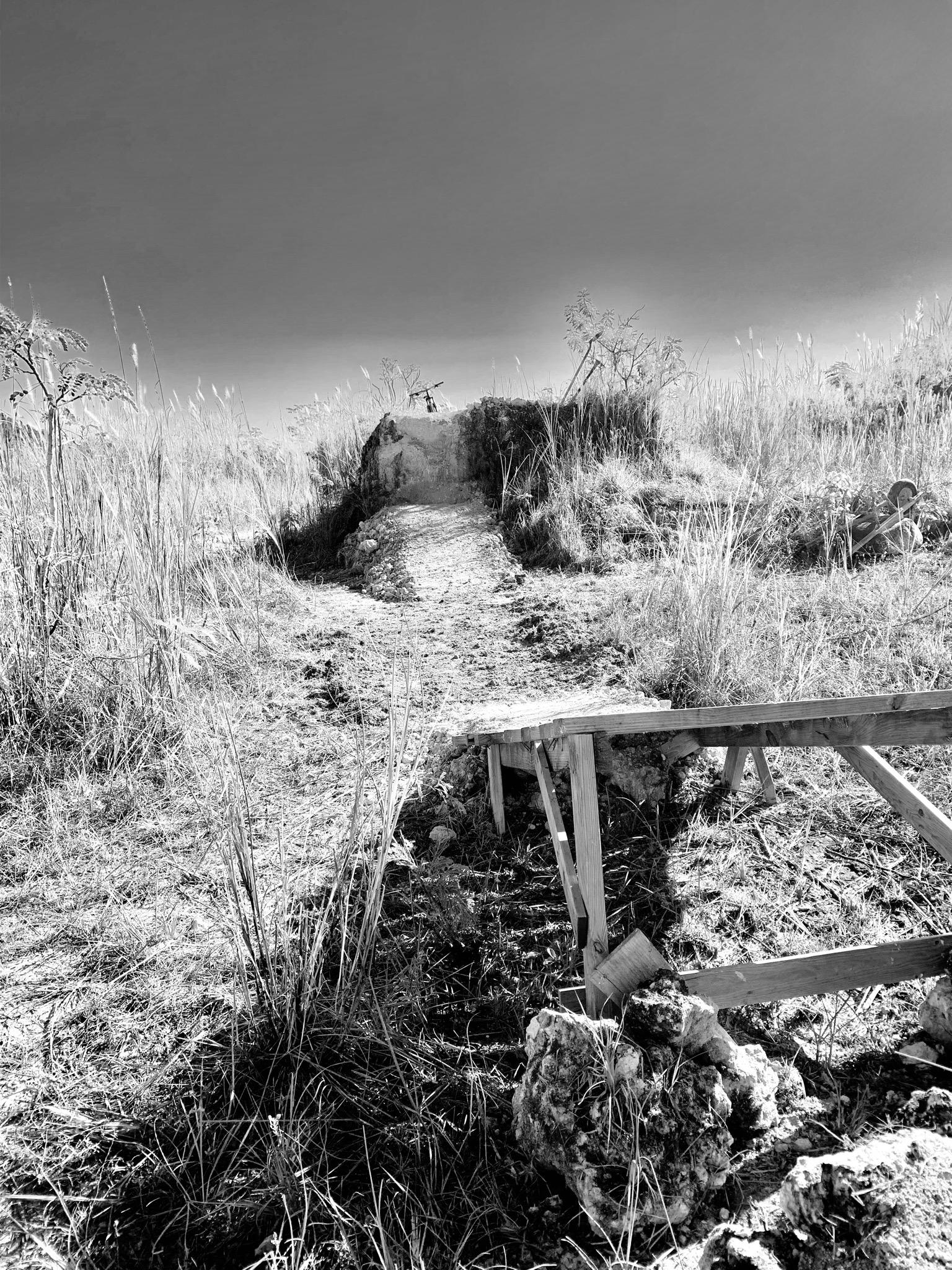 A black-and-white photograph depicting a narrow pathway leading through dry grass toward a small elevated mound. To the right, there is a makeshift wooden structure partially supported by stones, suggesting a rustic setting in a natural landscape. The sky above is clear, enhancing the contrast between the foreground and background elements. Monte Realengo mountain bike trail.