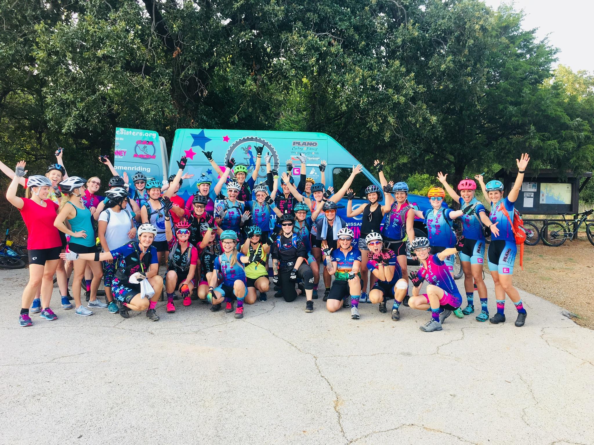 A large group of women wearing colorful cycling gear and helmets is gathered outdoors, smiling and cheering. They are positioned in front of a teal van adorned with vibrant graphics. Bicycles are parked nearby amidst a natural setting with trees in the background.
