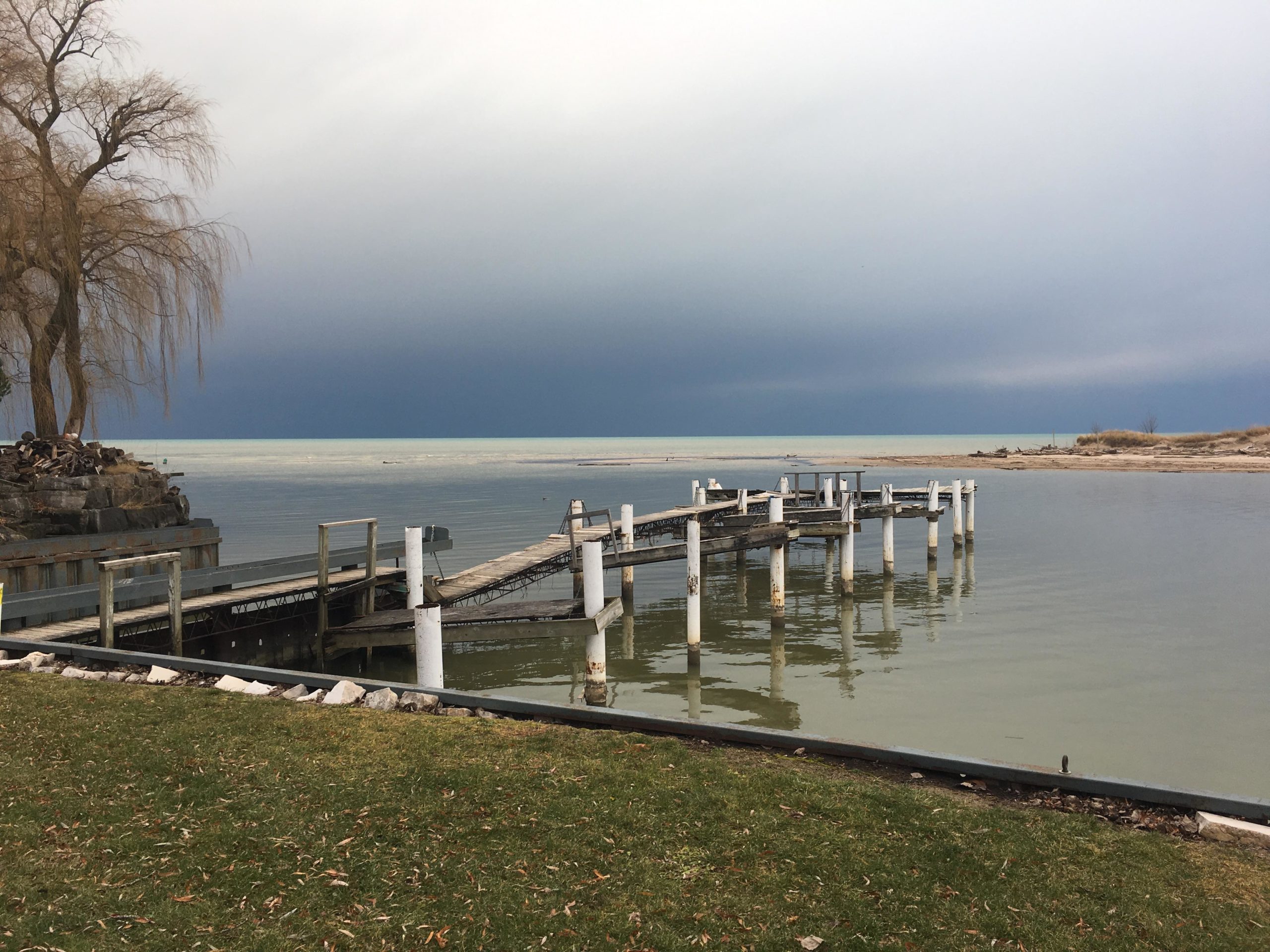 A calm lakeside scene featuring a wooden pier extending into the water. In the background, dark, overcast skies create a moody atmosphere, while a lone weeping willow tree stands near the shore. The water is still, reflecting the surroundings, and there are patches of sandy beach and grass on the shore. Lambton County Heritage Forest mountain bike trail.