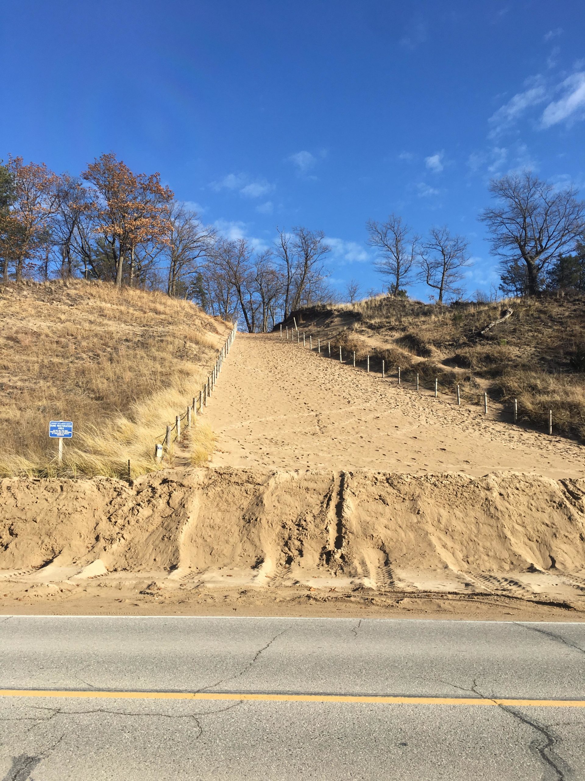 A sandy pathway leading up a hillside, flanked by grassy areas and sparse trees against a bright blue sky with scattered clouds. A sign indicating rules or information is visible at the base of the hill near the roadside. Lambton County Heritage Forest mountain bike trail.