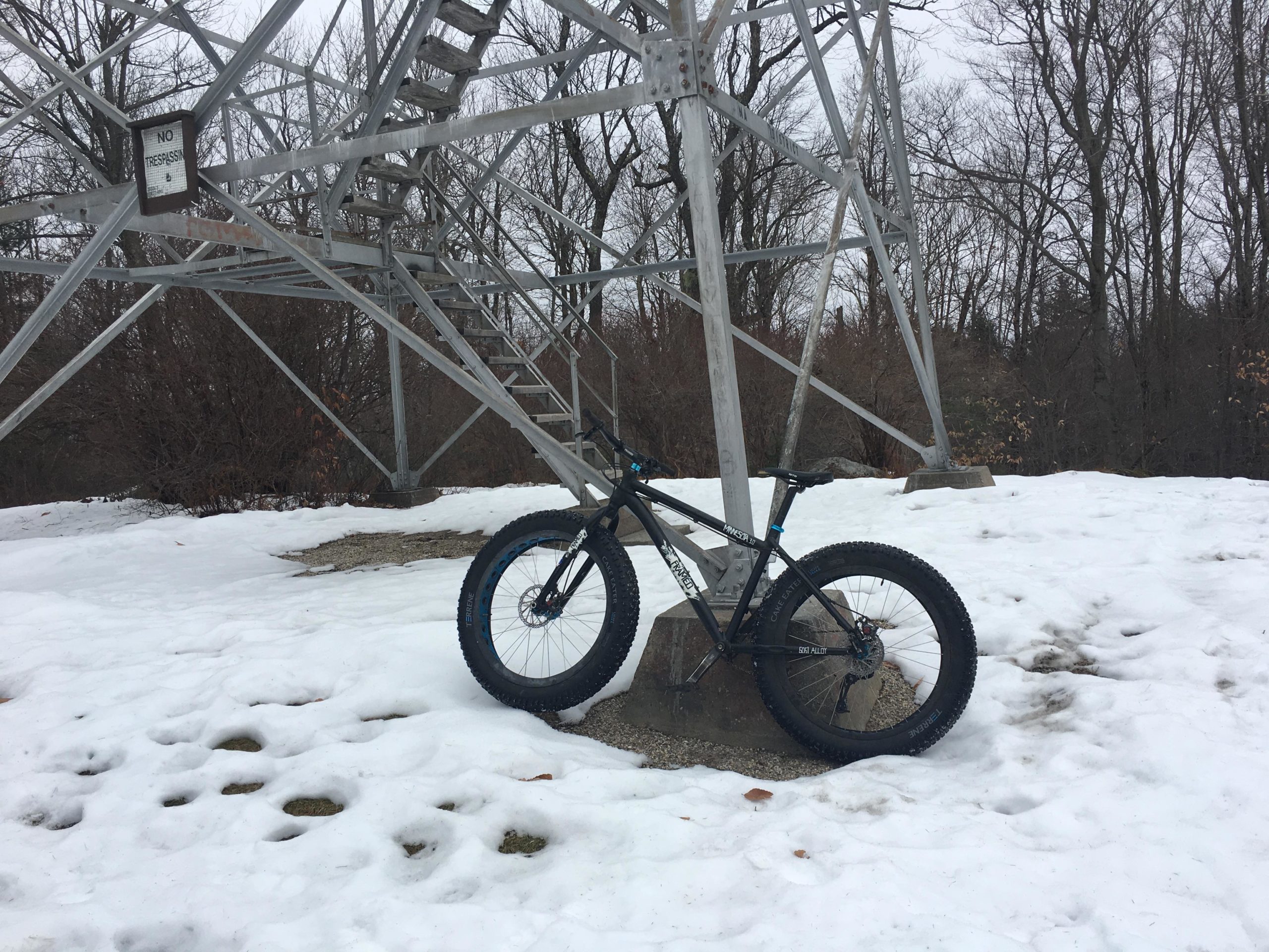 A fat tire bike parked on a snow-covered ground near a tall metal tower with a "No Trespassing" sign. Trees are visible in the background, and the scene is overcast, suggesting a chilly winter day. DAR mountain bike trail.