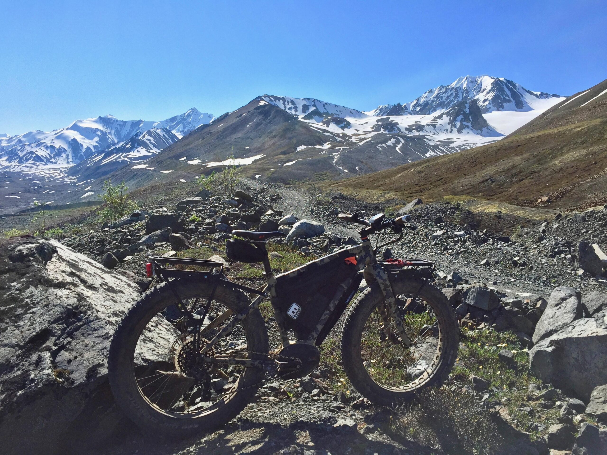 Felt Outfitter: A rugged fat bike positioned on a rocky trail with a backdrop of snow-capped mountains under a clear blue sky. The landscape features rolling hills and patches of greenery, showcasing a serene and adventurous outdoor setting.