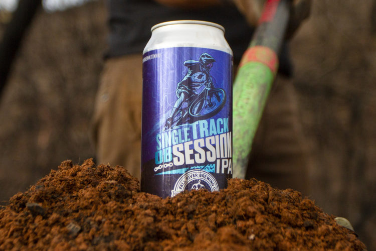 A can of "Single Track Session IPA" is positioned on a mound of dirt, with a blurred background showing a person holding a digging tool. The can features vibrant artwork of a cyclist and the brewery logo.