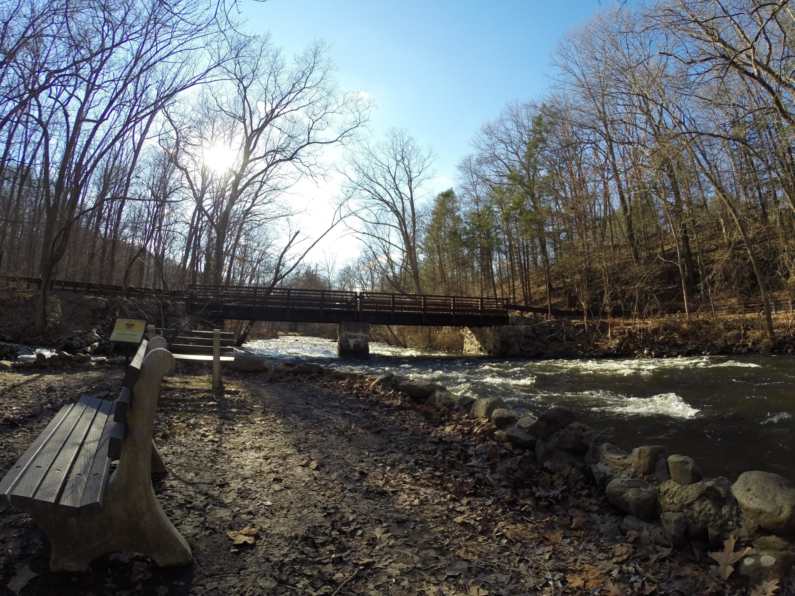 A tranquil stream flows through a wooded area, with a wooden bridge crossing above it. A stone bench is positioned nearby on the bank, surrounded by bare trees and a few remaining leaves. The sun shines brightly in the background, casting light on the scene. Allamuchy Mt. State Park: Deer Park mountain bike trail.
