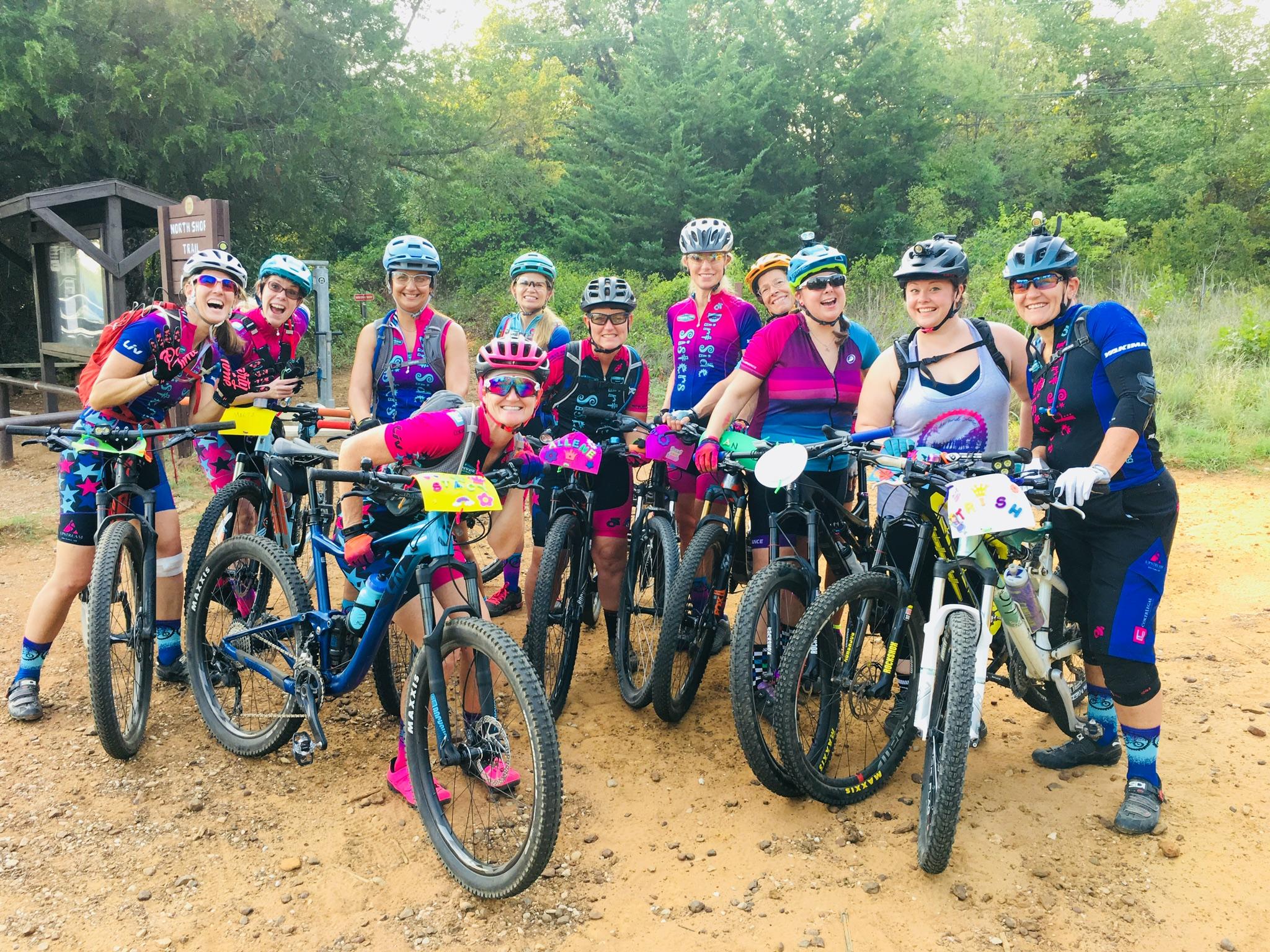 A group of women mountain bikers posing together with their bikes in a wooded area. They are dressed in colorful cycling gear and smiling, showcasing a sense of camaraderie and enthusiasm. The setting features dirt trails and greenery in the background.