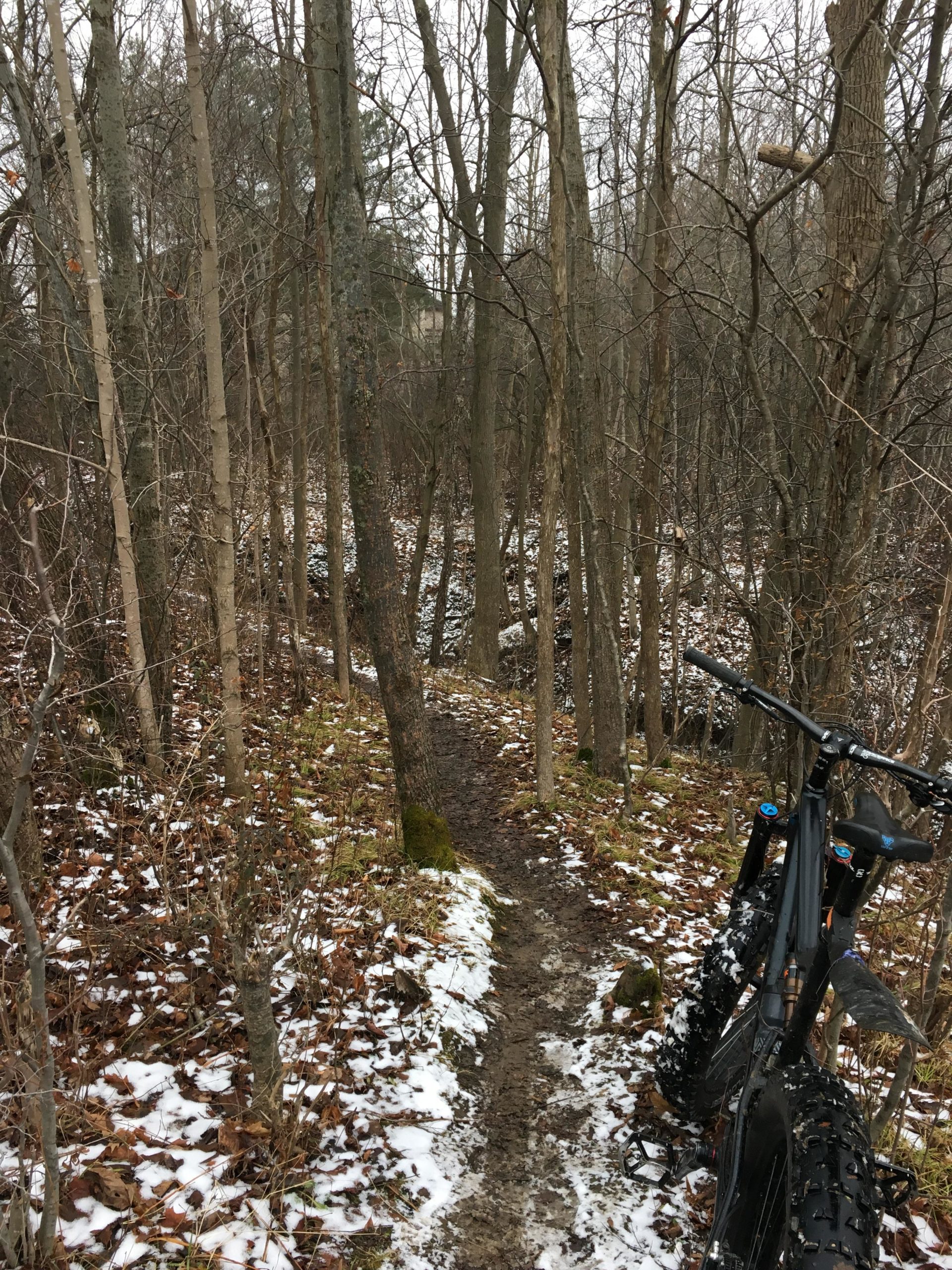 A narrow, muddy trail lined with bare trees and scattered snow, leading through a wooded area. In the foreground, a black mountain bike is parked to the right, partially obscured by the surrounding vegetation and snow. Western University trails mountain bike trail.