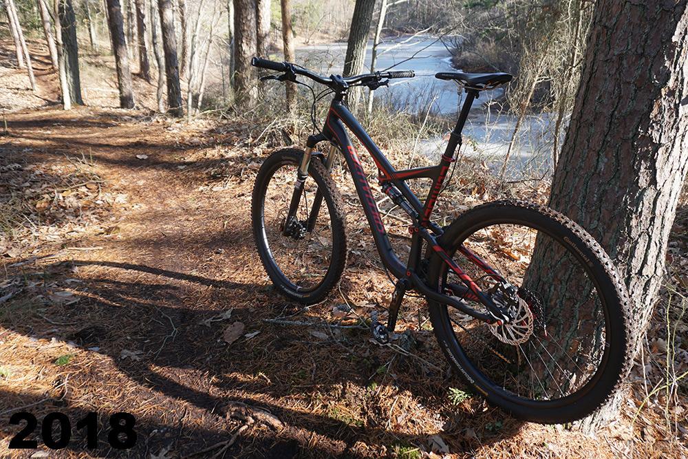 A mountain bike parked on a dirt trail surrounded by trees, with a river visible in the background. The scene features fallen leaves and a sunny atmosphere, suggesting an outdoor adventure. The year "2018" is displayed in bold text at the bottom left corner of the image. Maurice River Bluffs Nature Preserve mountain bike trail.
