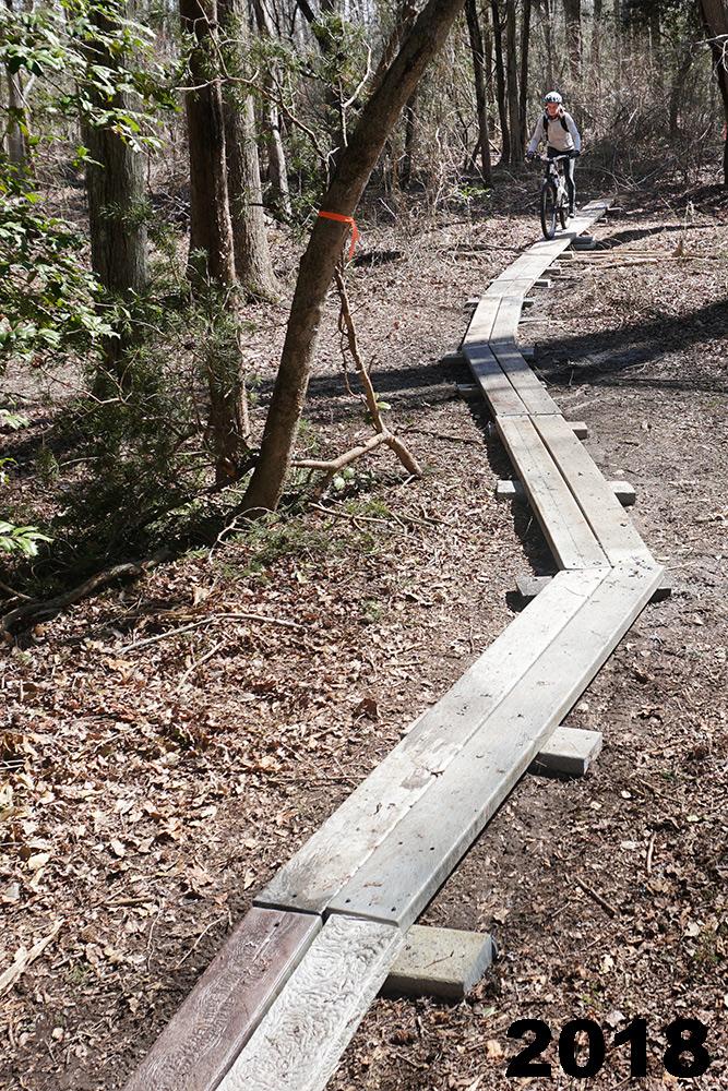 A mountain biker navigates a wooden boardwalk trail through a wooded area, surrounded by trees and dry leaves. The path curves gently, illustrating a natural landscape. The image is labeled "2018" in the bottom right corner. Maurice River Bluffs Nature Preserve mountain bike trail.
