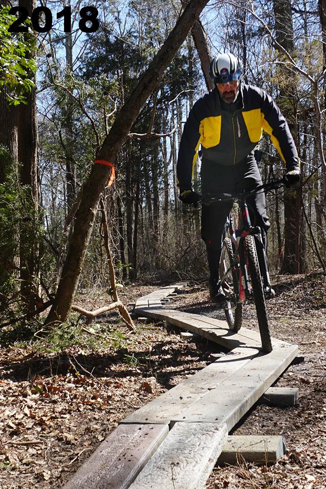 A cyclist navigating a narrow wooden plank trail through a wooded area, surrounded by trees and foliage. The cyclist is wearing a black and yellow jacket and a helmet, appearing focused as they ride. The image features the year "2018" prominently in the top left corner. The bluffs mountain bike trail.