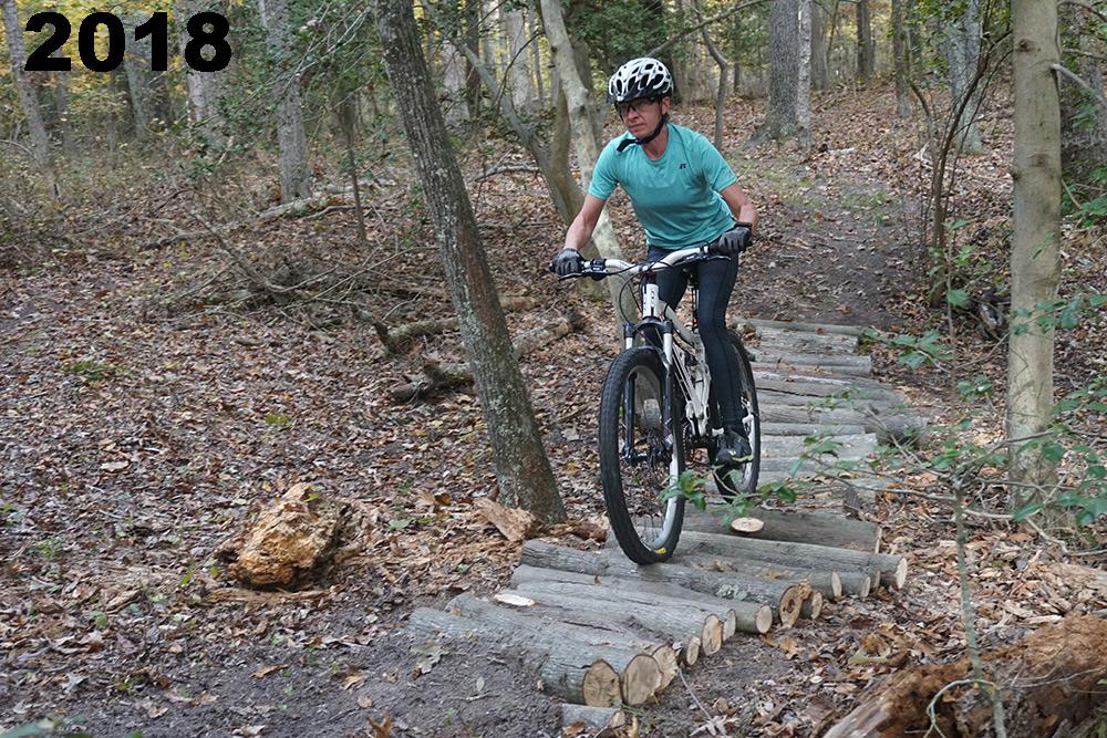 A person riding a mountain bike over a wooden bridge made of logs, navigating a trail in a wooded area during the fall. The scene includes trees, fallen leaves, and natural terrain, showcasing an outdoor biking experience. The year "2018" is displayed prominently in the top left corner. The bluffs mountain bike trail.
