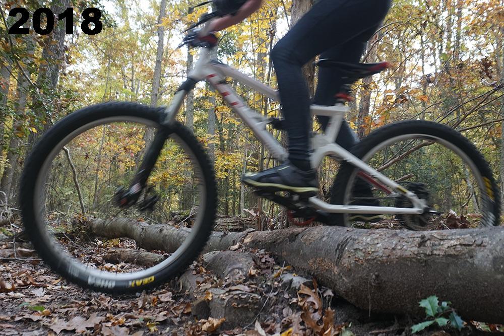 A mountain biker navigating over a fallen log on a trail surrounded by autumn foliage, with blurred motion conveying speed and movement. The year "2018" is prominently displayed in the top left corner. Maurice River Bluffs Nature Preserve mountain bike trail.