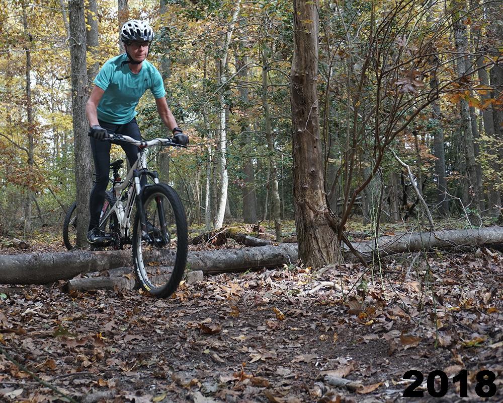A mountain biker navigating over a fallen log on a wooded trail during autumn. The cyclist is wearing a helmet and casual athletic clothing, surrounded by trees with colorful leaves. The ground is covered with fallen leaves and branches. The year "2018" is displayed in the bottom right corner. Maurice River Bluffs Nature Preserve mountain bike trail.