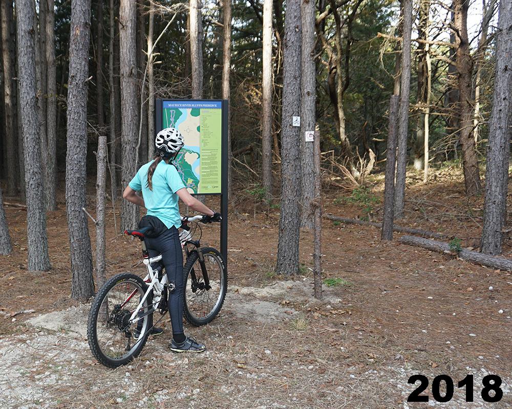 A person wearing a helmet and casual clothing stands next to a mountain bike, looking at a trail map sign in a wooded area. Tall trees and pine needles create a natural background. The image is labeled with the year "2018." Maurice River Bluffs Nature Preserve mountain bike trail.