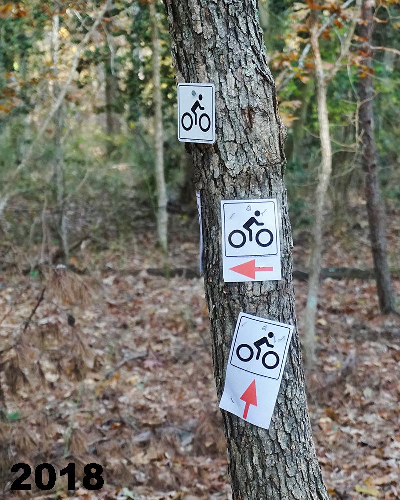 Three bicycle trail signs attached to a tree in a wooded area, indicating a biking path with arrows and bicycle symbols. The year "2018" is displayed in the bottom left corner. Maurice River Bluffs Nature Preserve mountain bike trail.