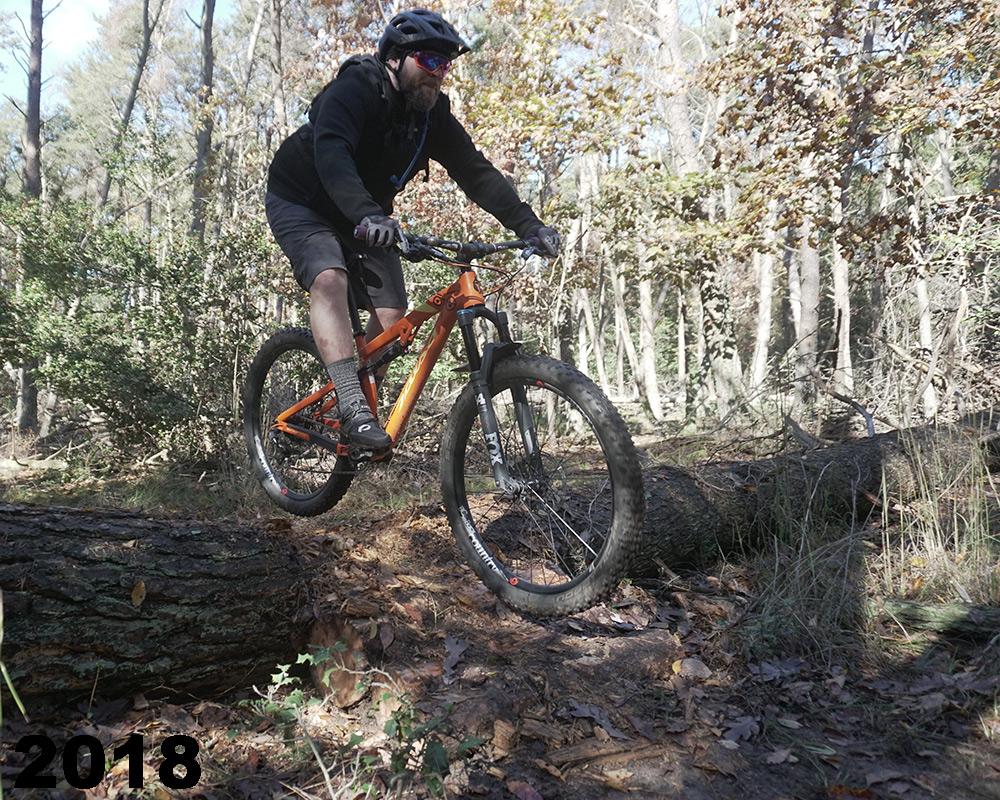 A mountain biker in a black hoodie and shorts jumps over a fallen log on a dirt trail in a forest, surrounded by trees with autumn leaves. The bike is orange, and the rider wears a helmet and sunglasses. The scene captures the excitement of mountain biking in nature. The year '2018' is displayed in the bottom left corner. Maurice River Bluffs Nature Preserve mountain bike trail.