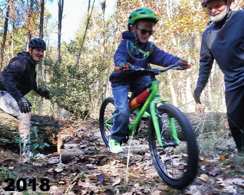 A young child rides a green bike on a dirt trail in a wooded area, with two adults nearby—one encouraging from the side and the other watching. The scene captures the joy of outdoor biking and the supportive atmosphere of a family activity. The image is set in a natural environment, surrounded by trees and fallen leaves. The year "2018" is displayed in the bottom left corner. Maurice River Bluffs Nature Preserve mountain bike trail.