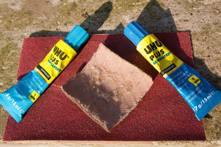 Two tubes of UHU Plus adhesive are placed on a textured surface, with one tube on either side of a square piece of sandpaper. The adhesive tubes have bright blue caps and yellow labels, displaying product information, while the sandpaper is red and shows signs of wear. The background is a neutral color.