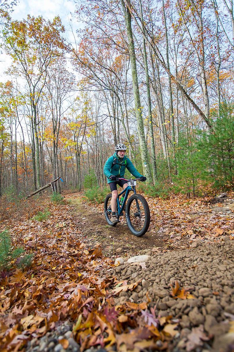 Trek farley 5: A person riding a mountain bike on a narrow dirt trail surrounded by trees with autumn foliage. The ground is covered in leaves, and there are patches of dirt and rocks along the trail. The rider is wearing a helmet and a blue jacket, showing an active outdoor lifestyle.