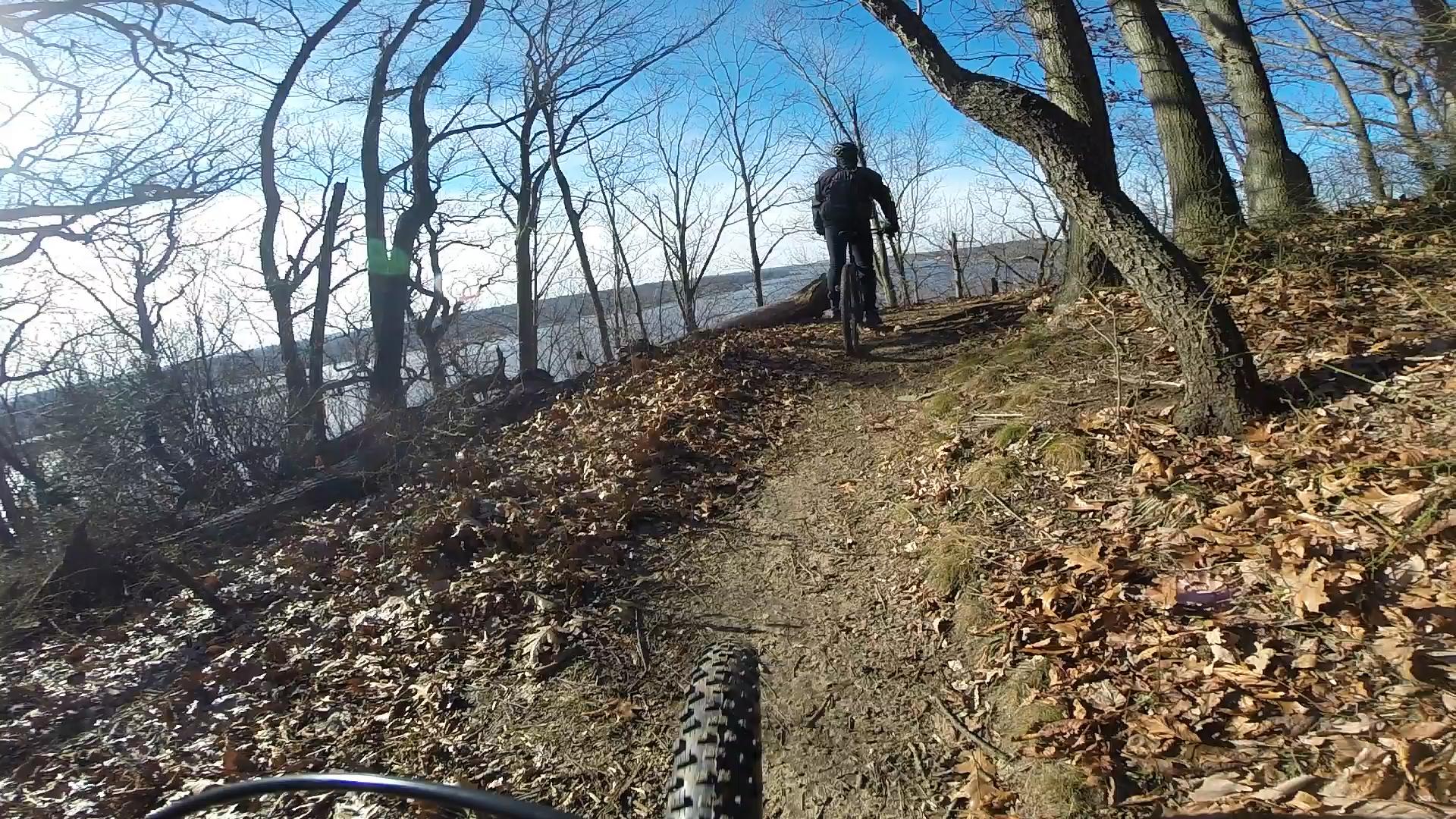 A mountain biker rides along a dirt path through a wooded area with bare trees and fallen leaves, with a lake visible in the background under a blue sky. Hartshorne Woods Park mountain bike trail.
