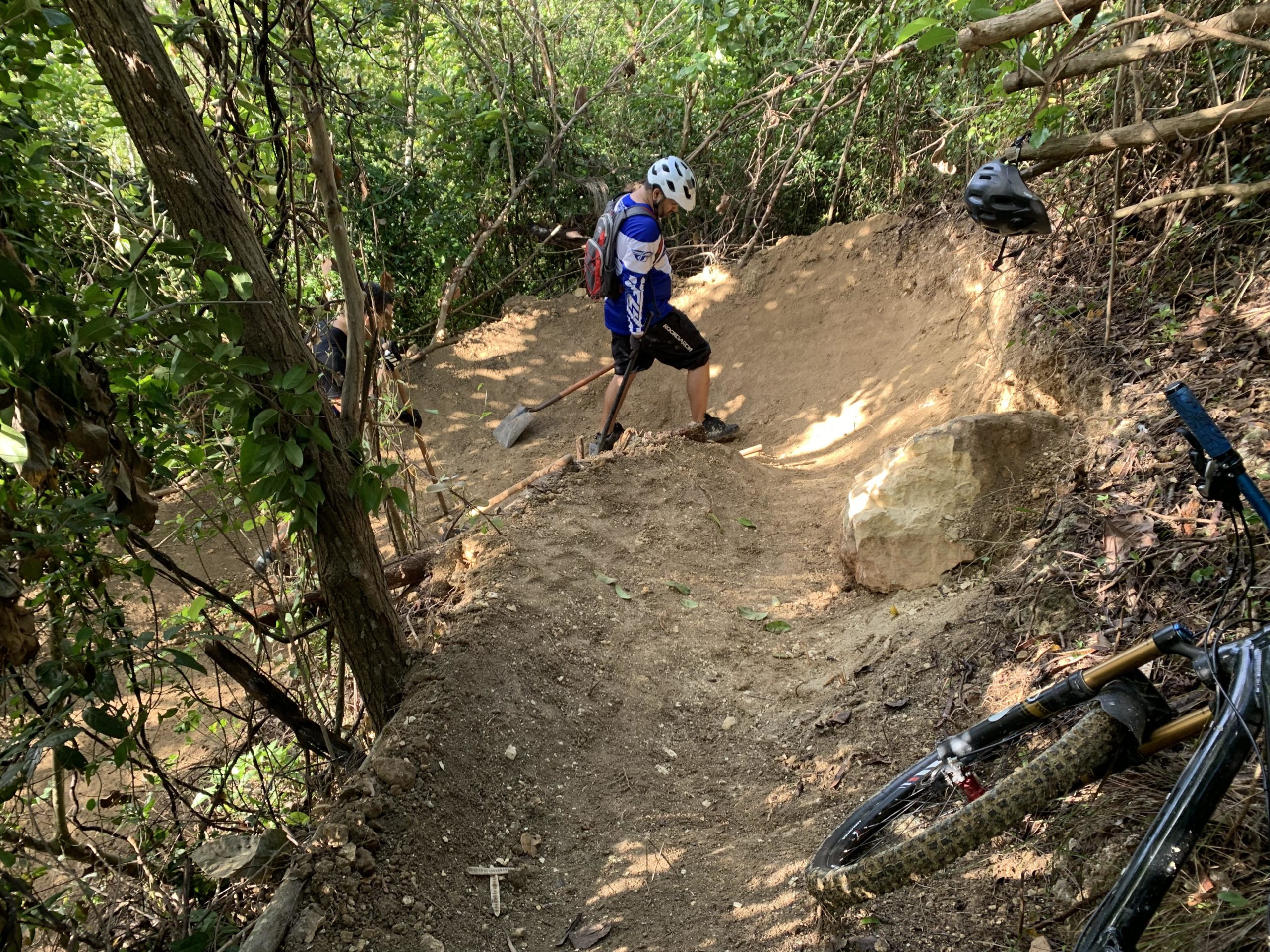 A mountain biker in a blue jersey and helmet is using a shovel to work on a dirt trail in a wooded area. Nearby, another person can be seen in the background also engaged in trail maintenance. A bicycle is resting on the ground to the side, surrounded by lush greenery and natural terrain. Monte Realengo mountain bike trail.
