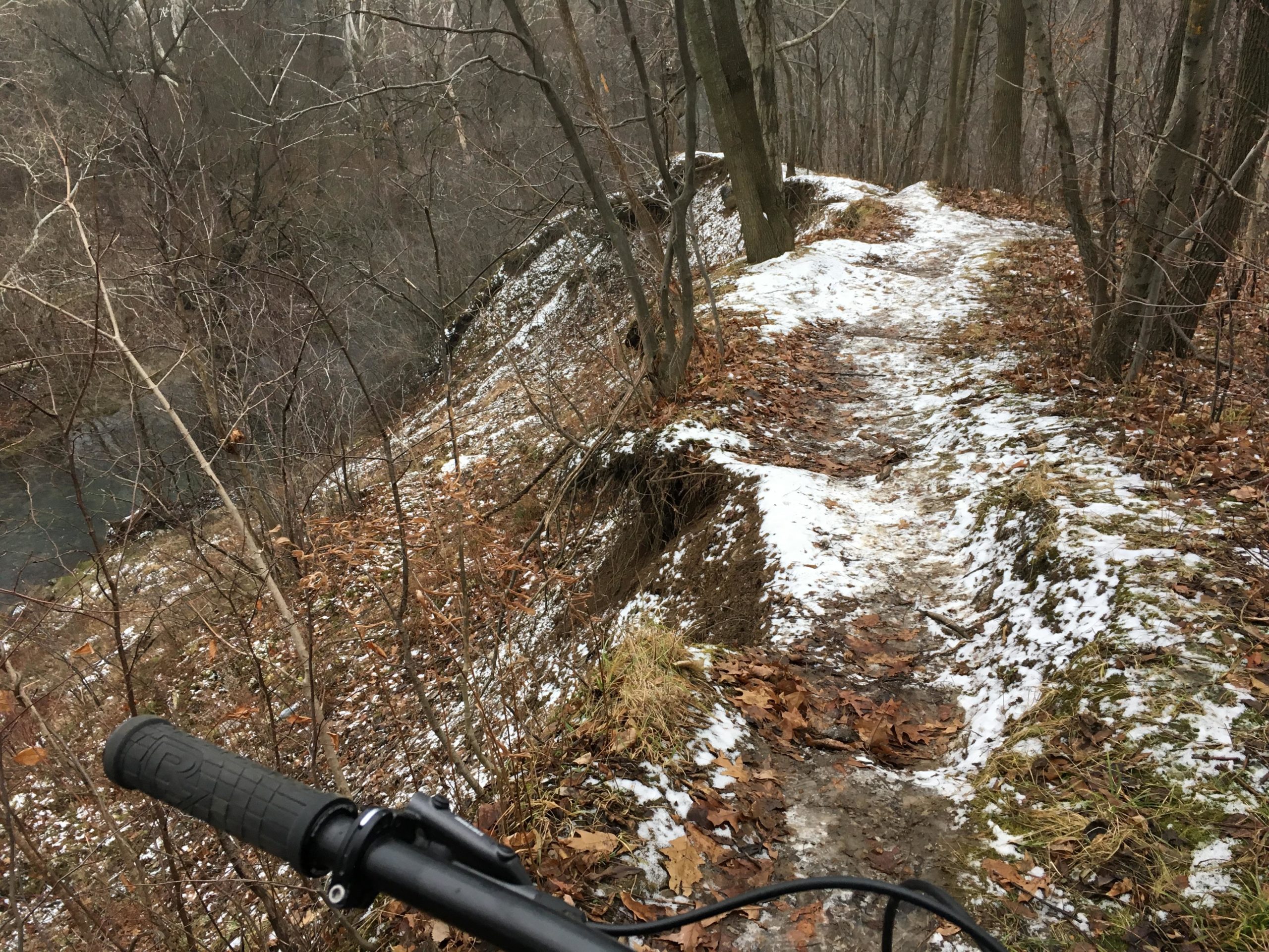 A narrow, winding trail through a forest, with snow and fallen leaves visible along the path. In the foreground, the handlebar of a mountain bike is partially visible, suggesting a biking perspective. The trail is bordered by trees and leads downhill towards a river at the bottom of a steep slope. Western University trails mountain bike trail.
