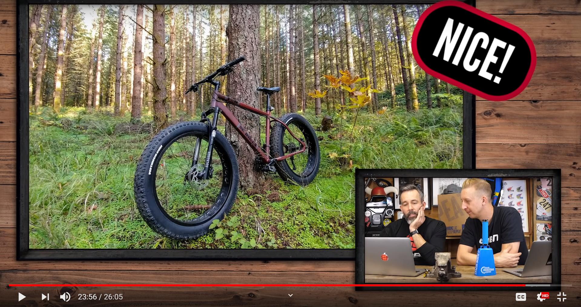 SE F@r: A mountain bike leaning against a tree in a lush forest setting, with tall trees and green foliage in the background. In the lower part of the image, two individuals are engaged in conversation, discussing biking-related topics, with laptops and equipment visible. A graphic displaying the word "NICE!" is overlayed in the corner.