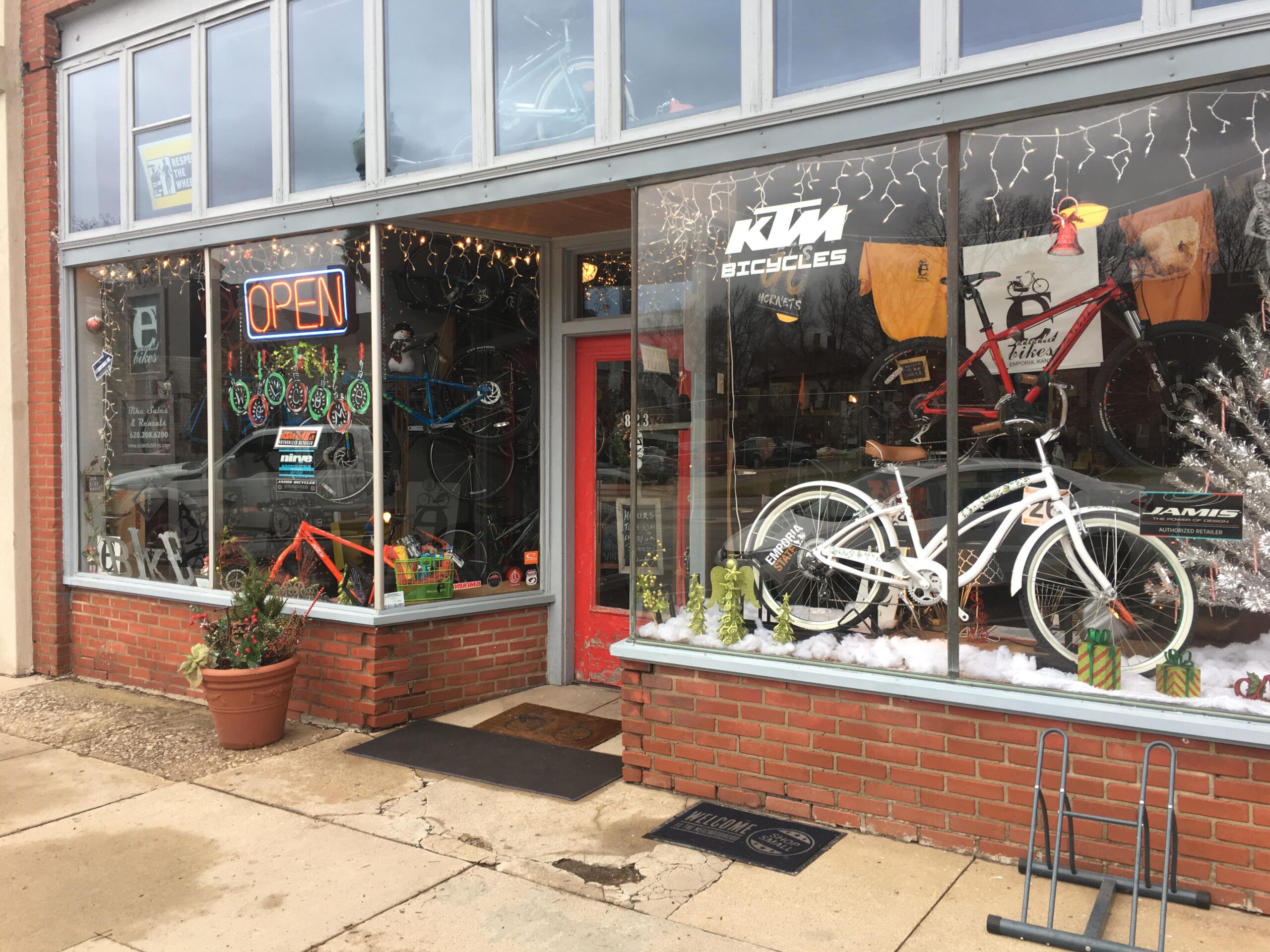 A view of a bicycle shop storefront featuring large windows showcasing various bicycles, including an orange and blue mountain bike and a white cruiser bike. The open sign is illuminated, and decorative lights hang in the window. There are potted plants near the entrance, and holiday-themed decorations are visible in the window display. A welcome mat is placed outside the door.