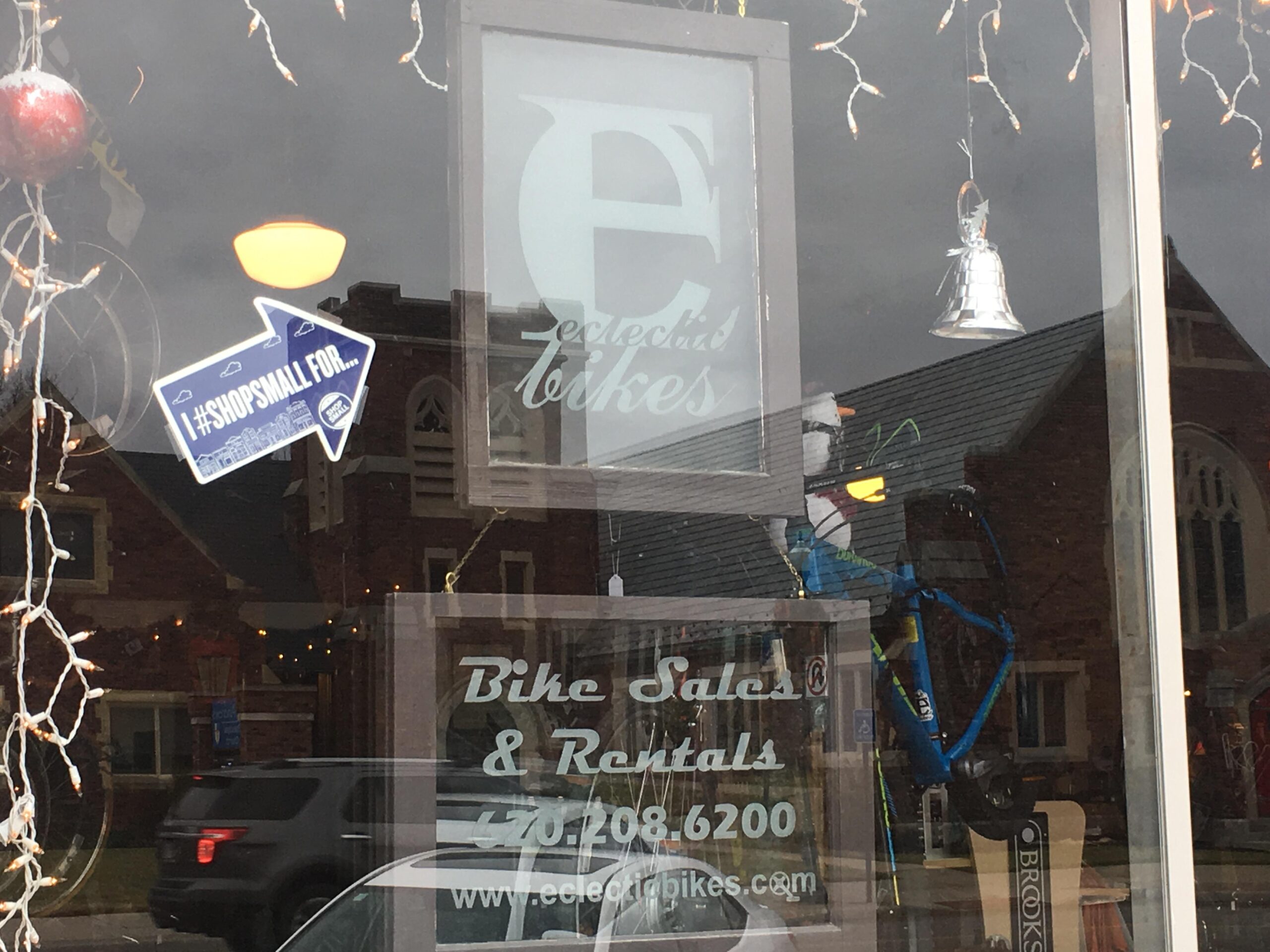 A close-up view of a bike shop window featuring signs that read "Eclectic Bikes," "Bike Sales & Rentals," and a hashtag promoting small businesses. The window display includes a silver bell decoration and a blue bicycle in the reflection, along with holiday lights and a festive atmosphere.