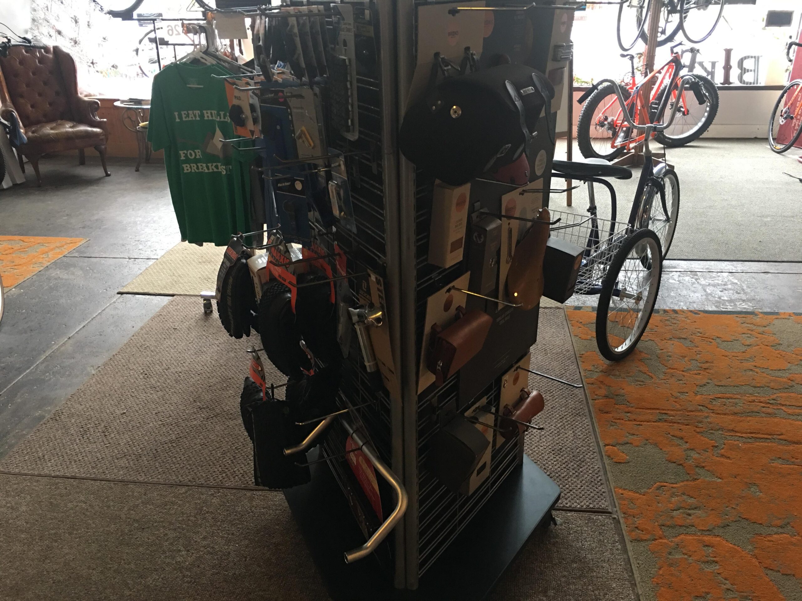 A display rack in a bike shop featuring various bicycle accessories, including gloves, bags, and tools. In the background, there is a green T-shirt with the text "I EAT HILLS FOR BREAKFAST," a brown leather chair, and several bicycles. The floor has a patterned area rug and a rubber mat near the entrance.