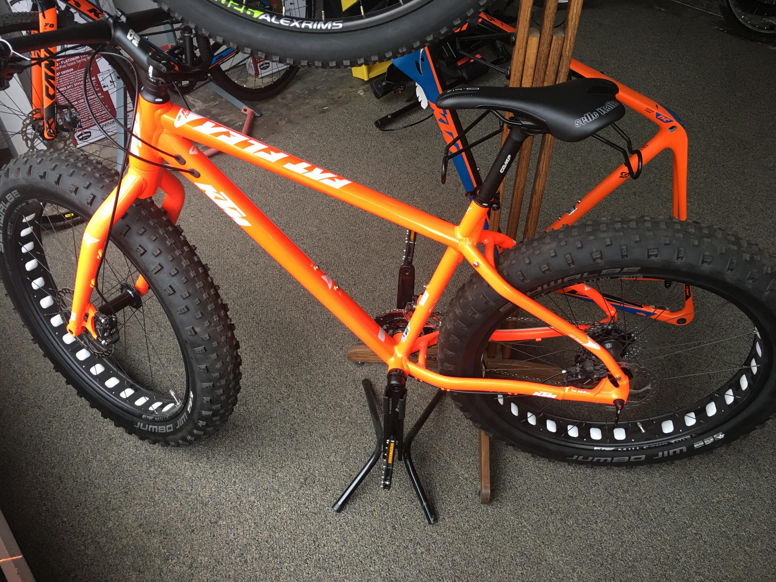 A bright orange fat tire bicycle is displayed in a bike shop. The bike features large, chunky tires and a sleek frame design. In the background, additional bicycles can be seen, highlighting a vibrant and active cycling environment.