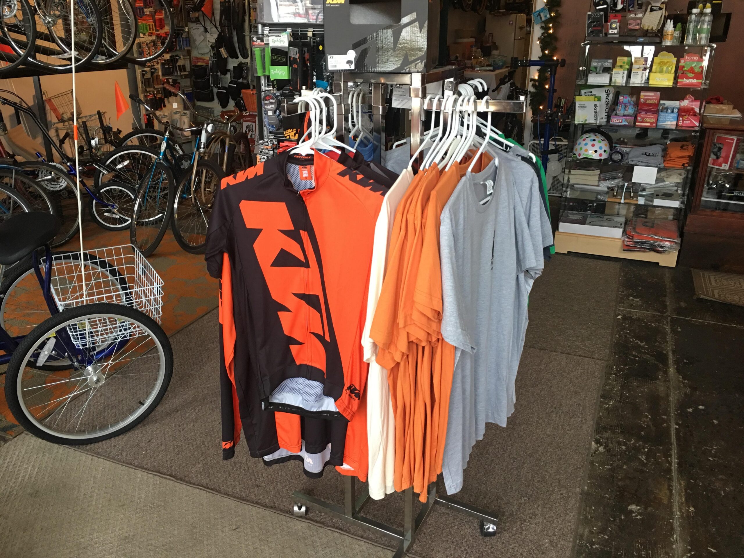 A display rack in a bicycle shop featuring cycling jerseys, including a black and orange KTM jersey, alongside a variety of casual t-shirts in shades of orange, gray, and white. In the background, bicycles and various cycling accessories can be seen.
