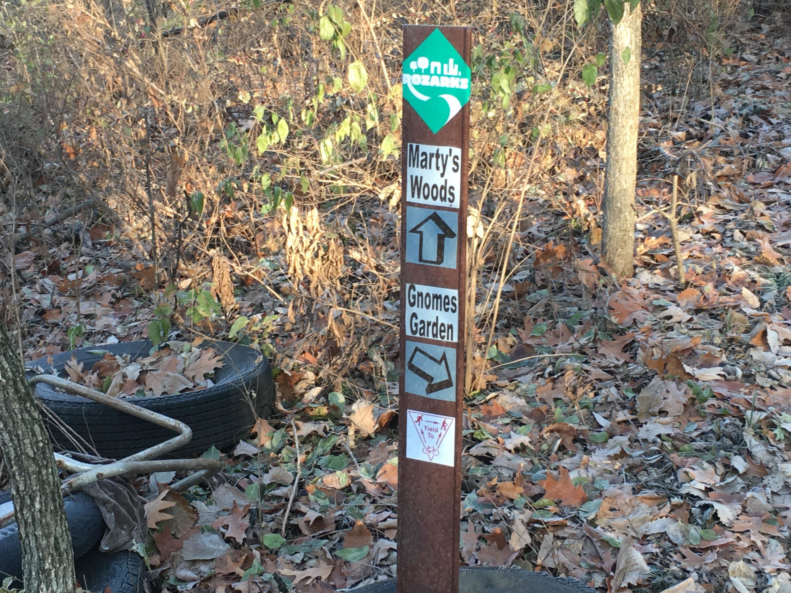 Signpost in a wooded area indicating trail directions: "Marty's Woods" points upward and "Gnomes Garden" points left. The ground is covered with fallen leaves, and there are old tires and a metal frame nearby. Rozarks mountain bike trail.