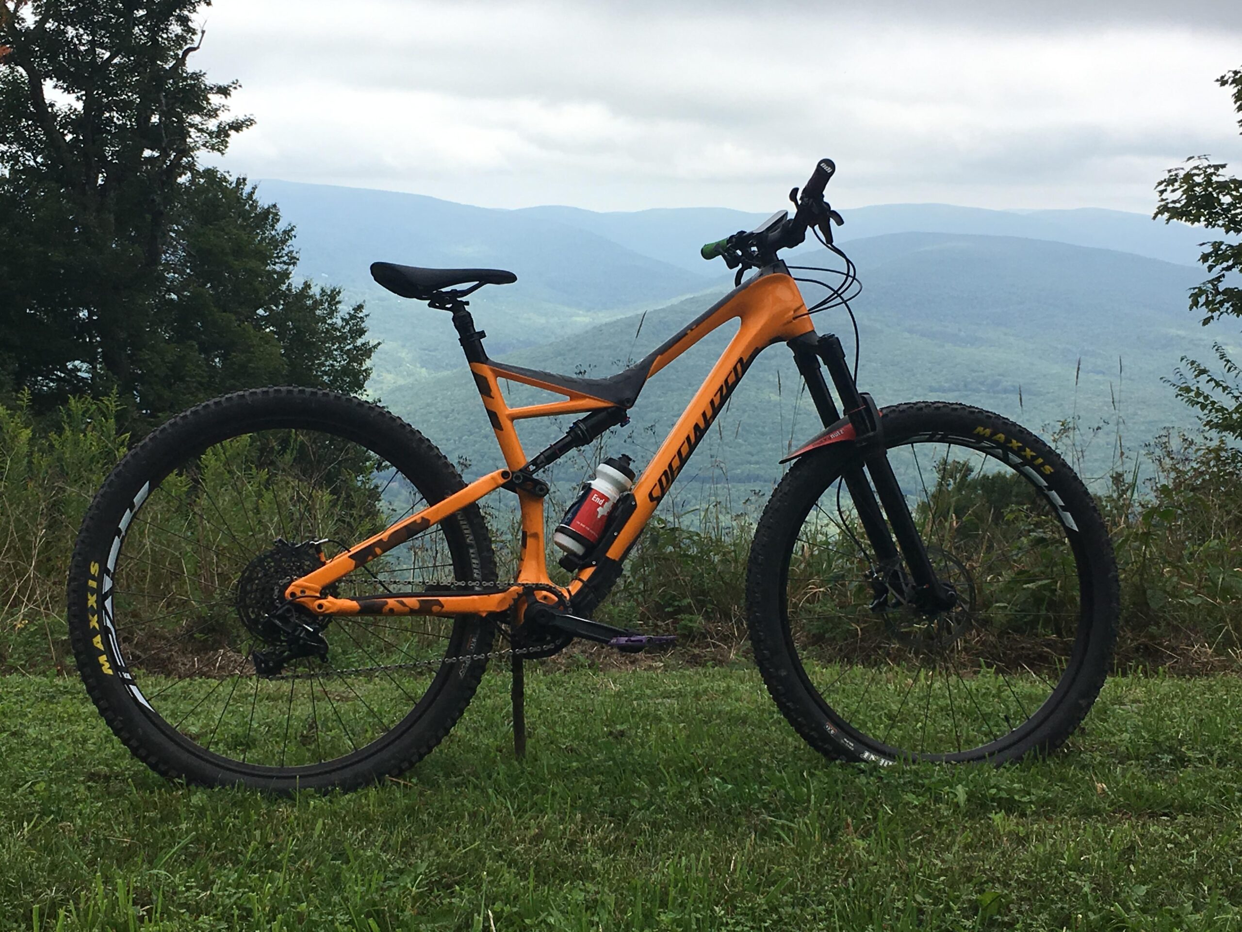 Specialized Stumpjumper Comp Carbon 29: A bright orange mountain bike stands on a grassy area, with a scenic view of rolling mountains in the background under a cloudy sky. The bike features large tires and a water bottle attached to its frame, showcasing an outdoor adventure setting.