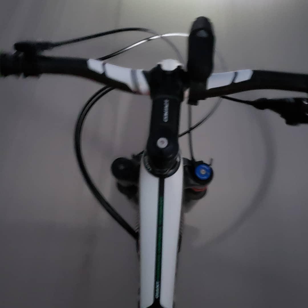 Image of a bicycle's handlebars and front fork, viewed from above, with a dimly lit background. The handlebars have a white and black design, along with brake and gear shifters. The image shows part of the bike's frame and some components related to suspension near the front wheel. Virginia Key North Point mountain bike trail.