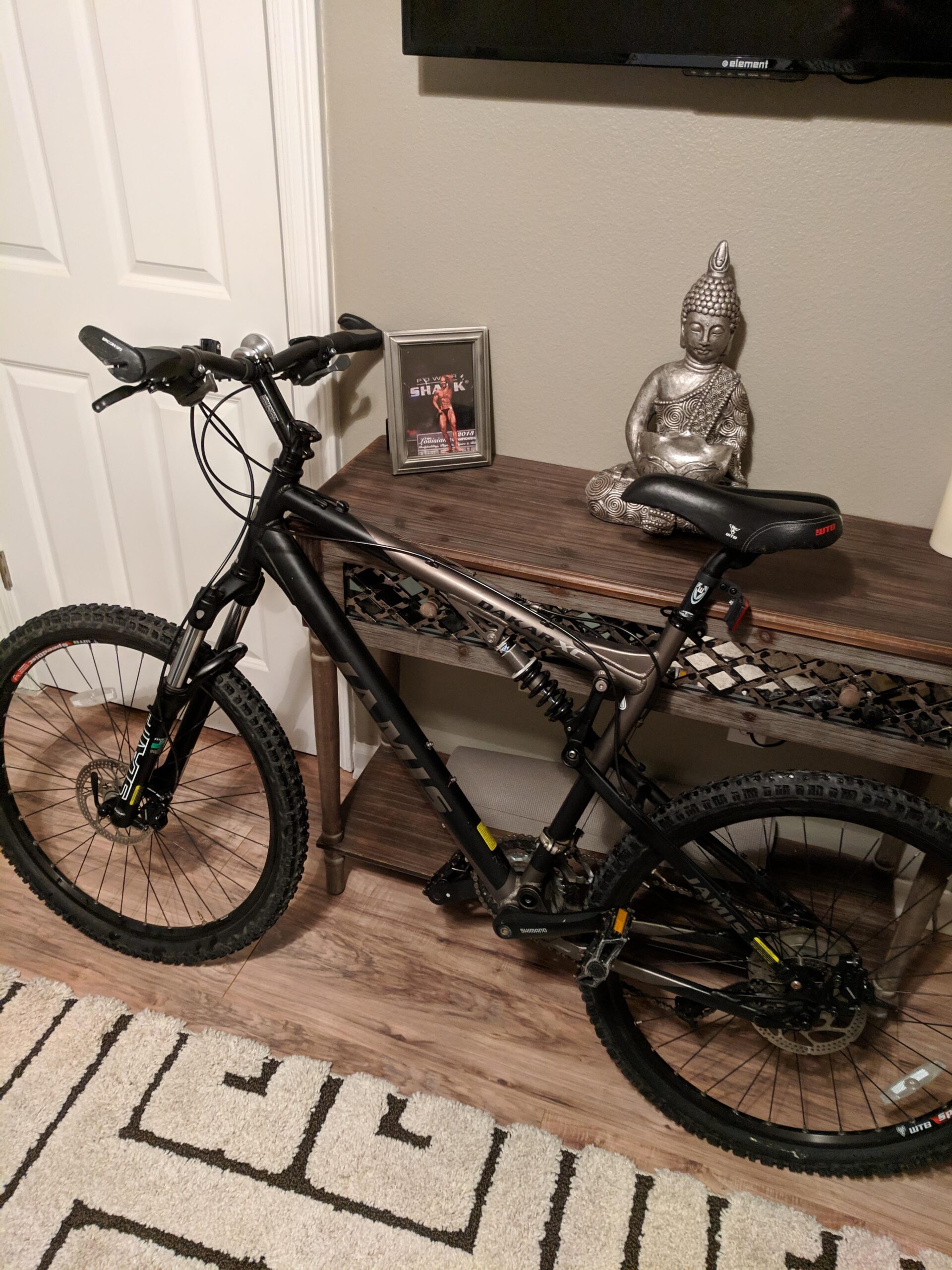 Jamis Dakar XC: A black mountain bike leaning against a wooden console table, next to a decorative Buddha statue and a framed picture. The bike features thick tires and a suspension system. The background includes a light-colored wall and a TV mounted above the table. A patterned rug is visible on the floor.