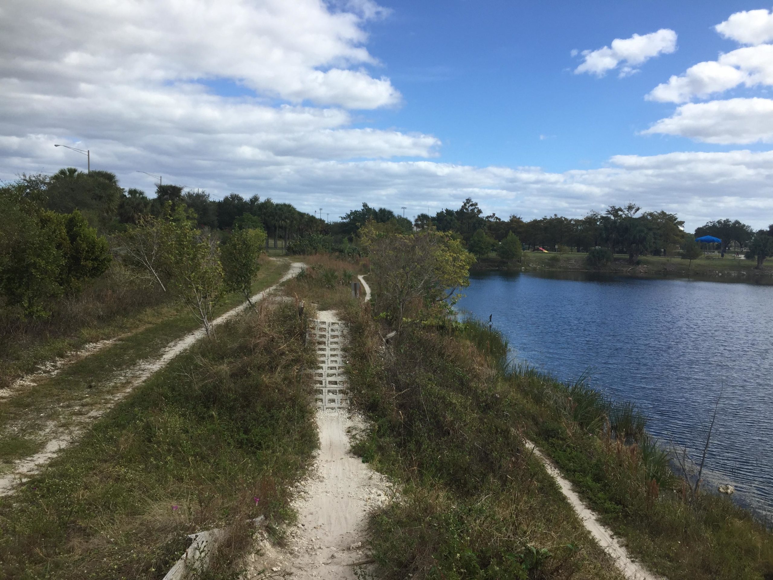 A scenic view of a pathway winding alongside a peaceful lake, surrounded by lush greenery and trees. The sky is partly cloudy, with a hint of blue peeking through. In the distance, there are some park facilities, including a shelter. The trails appear well-defined, inviting visitors to enjoy a leisurely stroll or bike ride. Quiet Waters Park mountain bike trail.