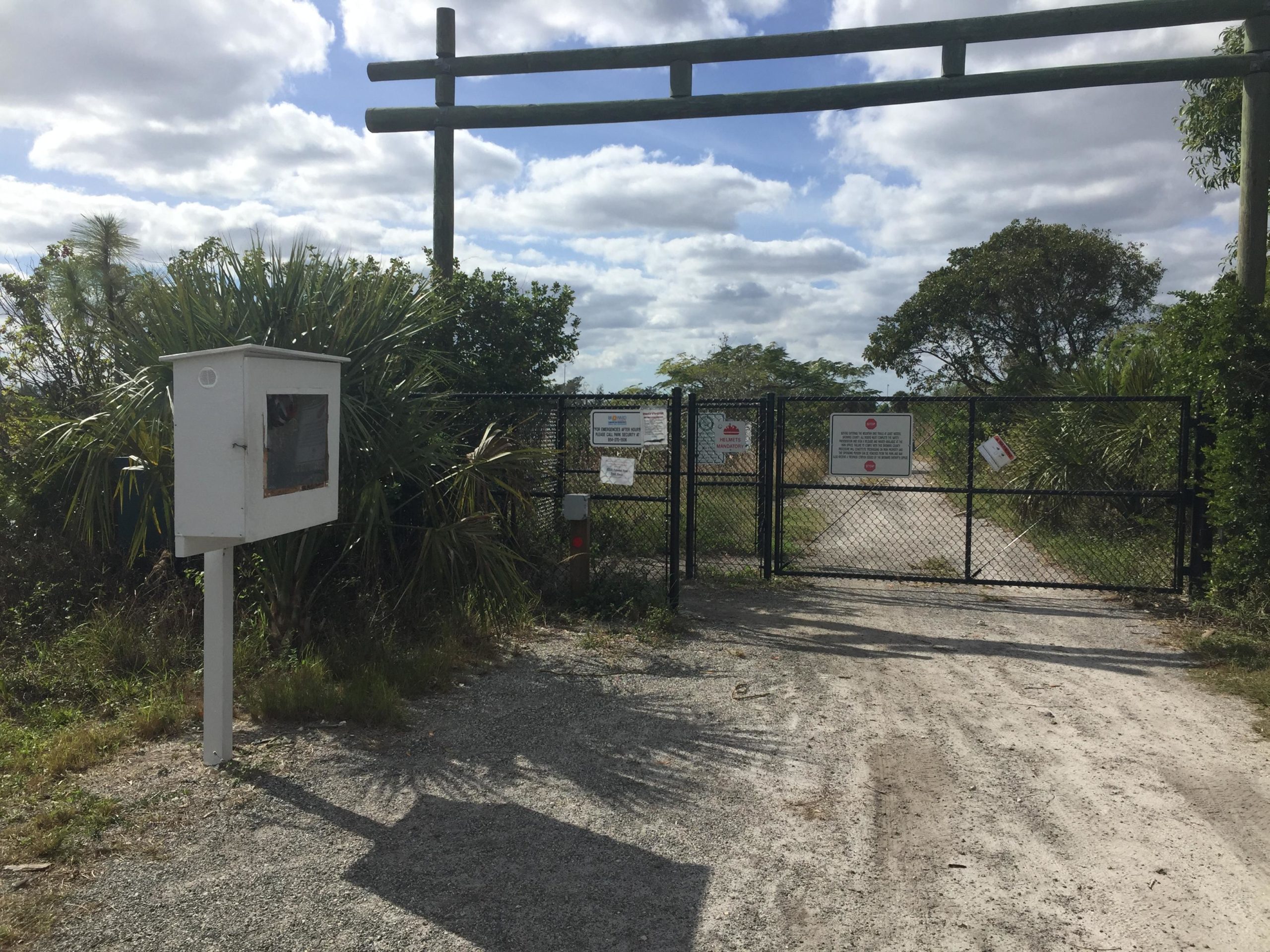 Alt text: A chain-link gate blocking a dirt pathway, flanked by green foliage and a wooden structure overhead. To the left, there is a small white information box on a post. Several signs are posted on the gate, indicating restrictions or rules. The sky above is partly cloudy. Quiet Waters Park mountain bike trail.